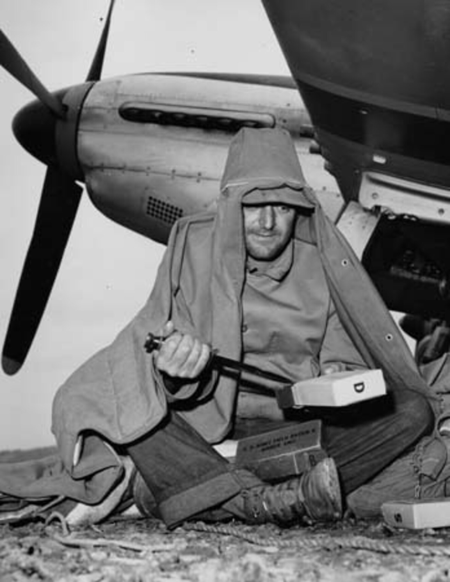 Huddled under a canopy blanket beneath the Wing of his 7th Air Force P-51 fighter on Iwo Jima, Crew Chief S/Sgt. Anthony A. Belesi, cuts open dry K-rations. Mechanics remained on the flight line 14 hours from dawn to dark with no shelter.