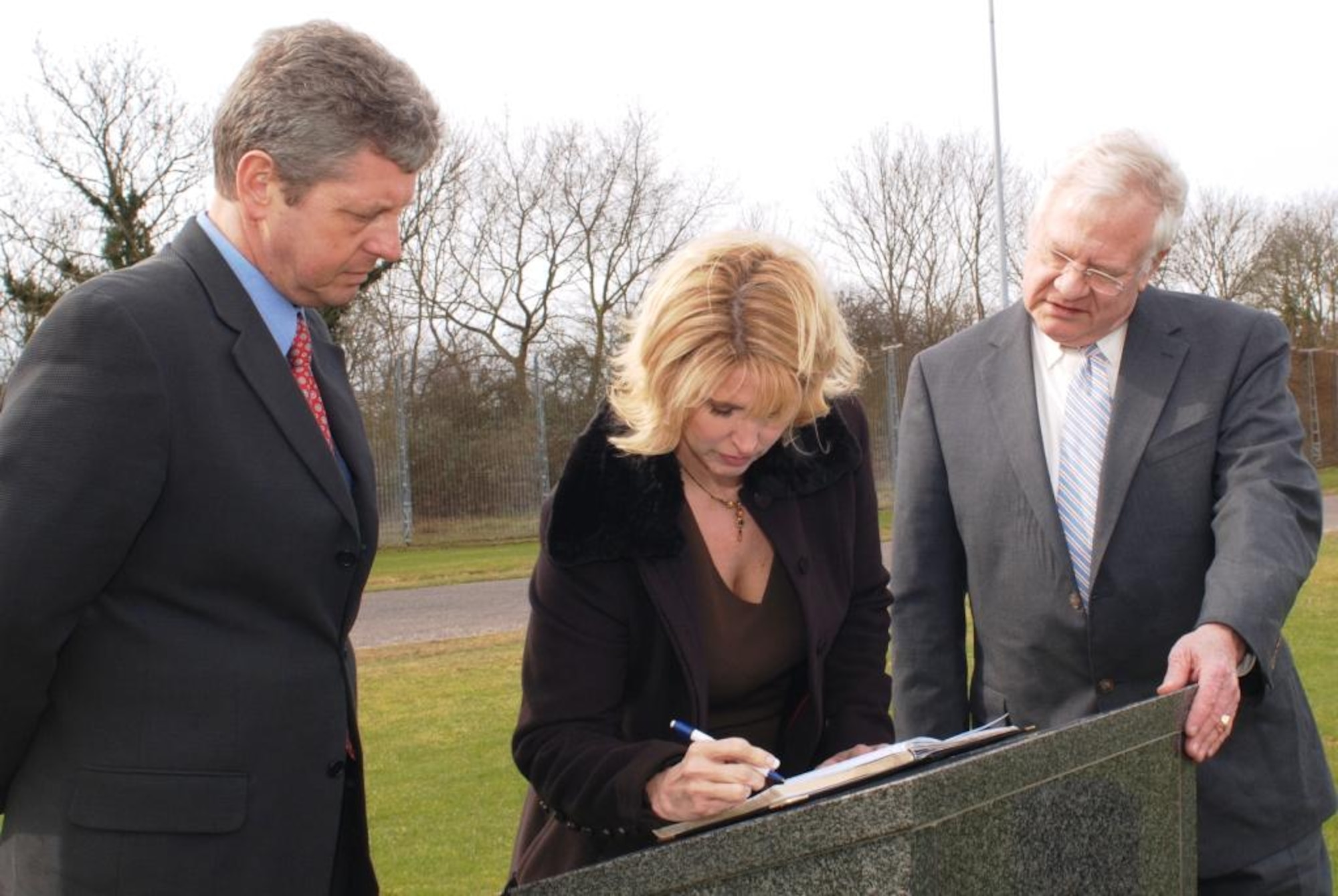 Ms. Suzanne Wilson signs the guest book at the memorial to the 303rd Bomb Group at RAF Molesworth, England as her partner Mr. Colin Poole (on her right) and Mr. Peter Park of the JAC Commander's Action Group look on.  Ms. Wilson represented her father, retired Lt Col Edgar Miller who flew as a B-17G Flying Fortress pilot from Molesworth during World War II, at a ceremony on March 13th.  (U.S. Air Force Photo by Staff Sgt. Jocelyn L. Rich)