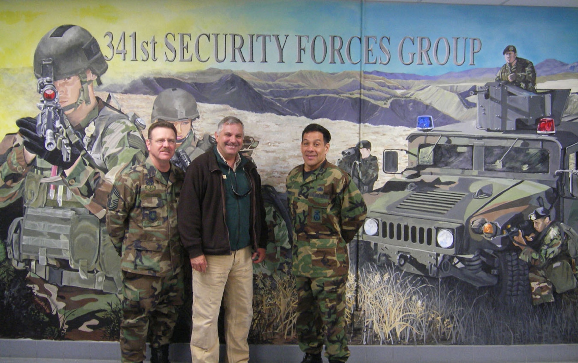 Left to right, Chief Master Sgt. Larry Wilson, 341st Security Forces Group manager; Mark Schaefer, BLM park ranger; and Col. Steve Asher, 341st SFG commander, pose in front of the wall mural in the building 500 basement – home of the 341st SFG. (U.S. Air Force photo)