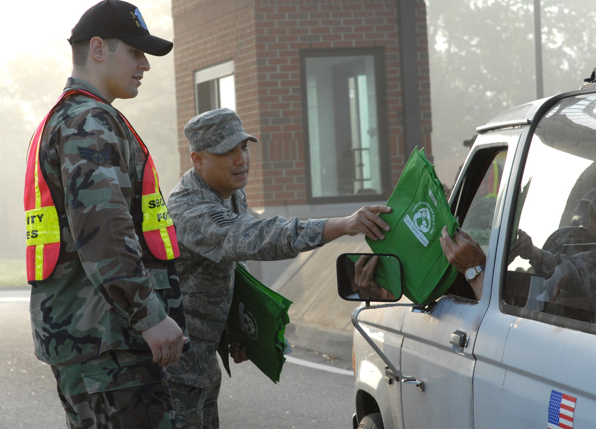 LANGLEY AIR FORCE BASE, Va. -- Tech. Sgt. Allan Araneta (left), 1st Logistics Readiness Squadron, hands out reusable grocery bags at the West Gate April 24 to show the Langley’s support of carpooling during earth week while Senior Airman Justin Cyr, a 1st Security Forces augmentee from the 30th Intelligence Squadron, checks identification. (U.S. Air Force photo/Senior Airman Christopher L. Ingersoll)