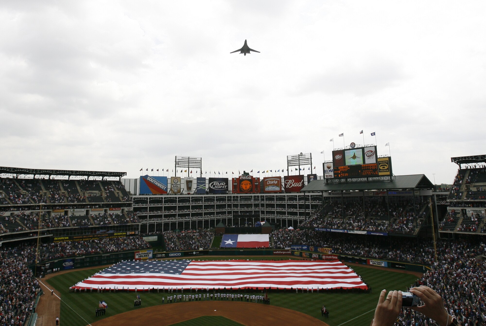 A B-1 Bomber from Dyess Air Force Base, Texas, flies overhead as nearly 50 Air Force Reserve and Guard members assist in revealing the 300-foot American flag during the Texas Ranger's Opening Day festitives. Many of the Reservists are stationed at the Naval Air Station Joint Reserve Base Carswell Field, Texas with the 301st Fighter Wing. (Courtesy Photo)