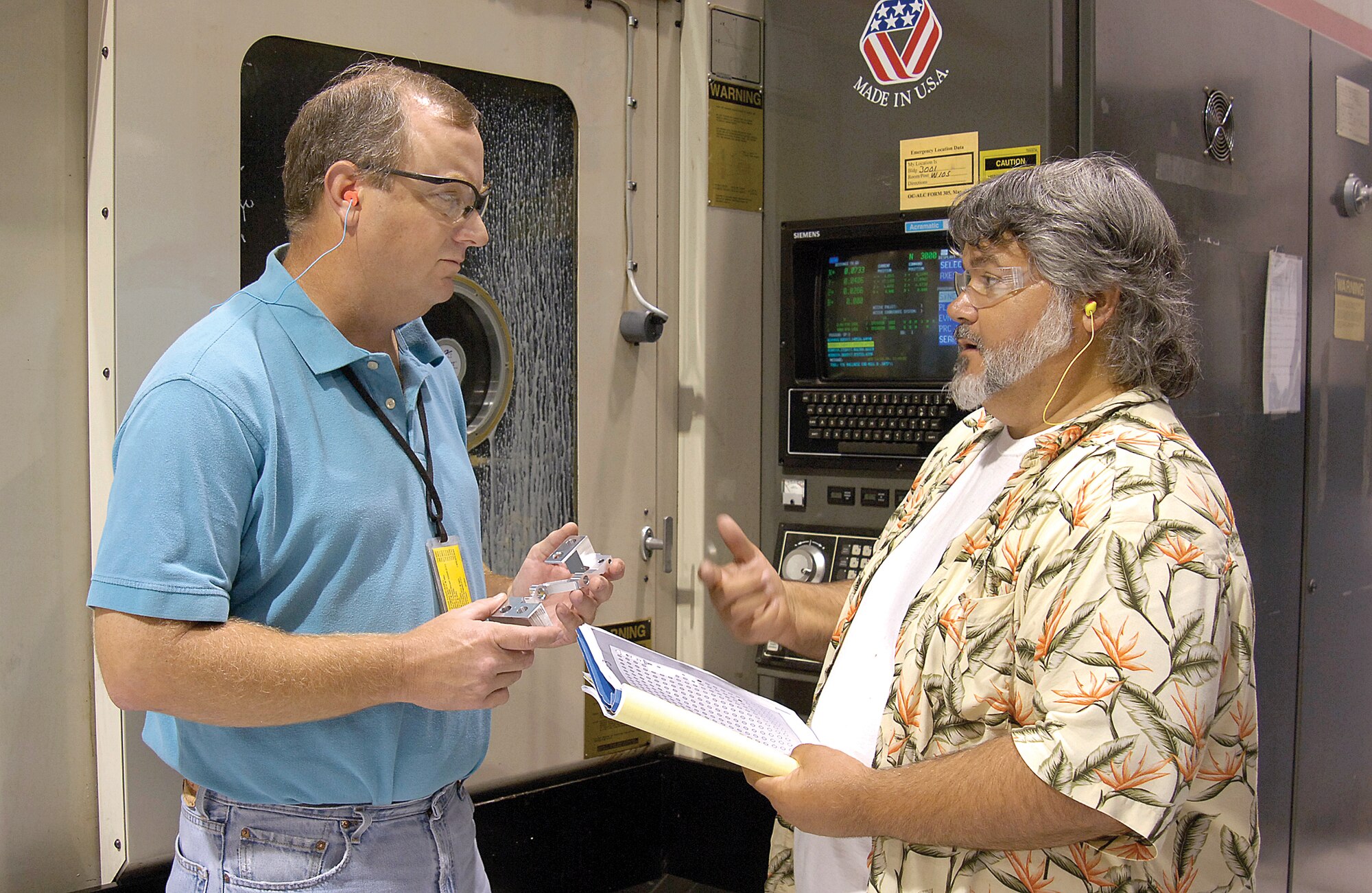 While a milling machine behind them precisely carves a T-38 left aileron actuator lever from raw material, Numerical Control programmer John Wilbanks, left, and machinist Michael Barnes discuss the process.  Oklahoma City Air Logistics Center’s 76th Maintenance Wing professionals joined other centers in a round-the-clock push to make hundreds of levers, helping Air Force officials keep the T-38 training jet flying.(Air Force photo/Margo Wright)