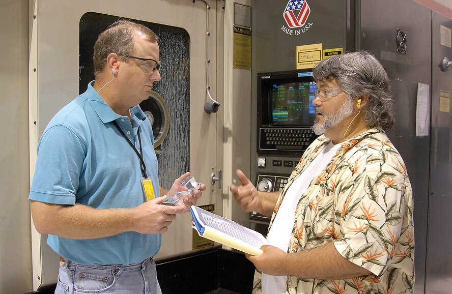 Numerical control programmer John Wilbanks (left) and machinist Michael Barnes discuss the T-38 project while a milling machine behind them precisely carves a T-38 left aileron actuator lever from raw material. (Air Force photo by Margo Wright)