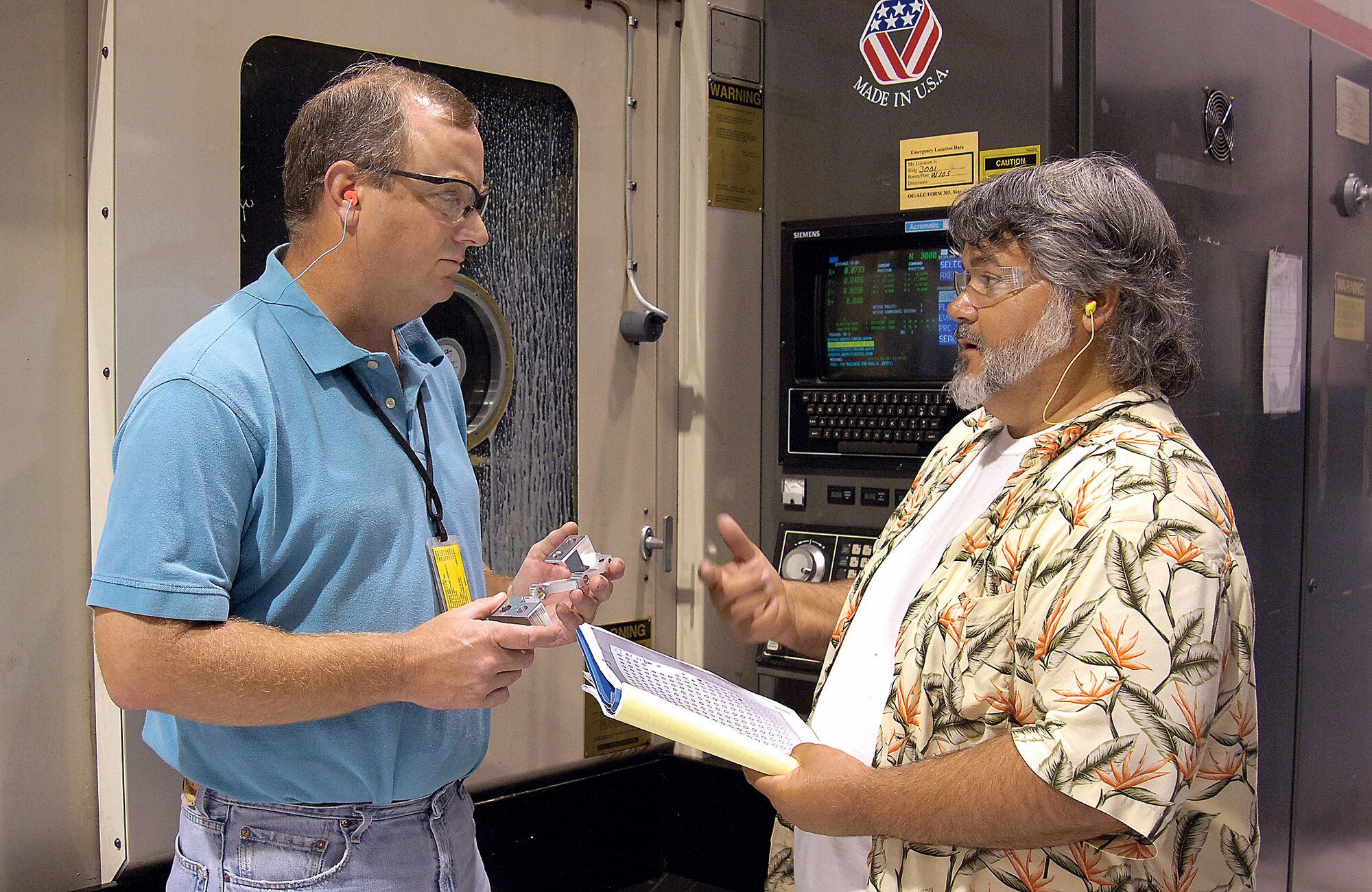 Numerical control programmer John Wilbanks (left) and machinist Michael Barnes discuss the T-38 Talon project while a milling machine behind them precisely carves a T-38 left aileron actuator lever from raw material at Tinker Air Force Base, Okla. (U.S. Air Force photo/Margo Wright) 
