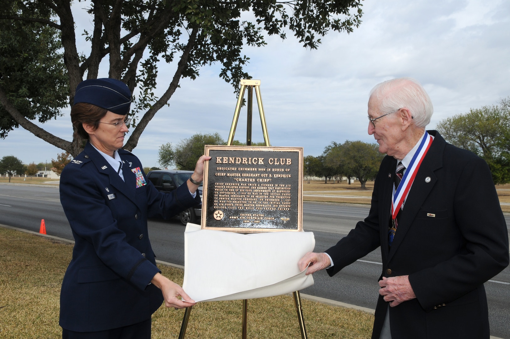 Col. Jacqueline Van Ovost, 12th Flying Traning Wing commander, and retired Chief Master Sgt. Guy Kendrick unveil the plaque redesignating the Randolph Air Force Base Enlisted Club as the "Kendrick Club." (U.S. Air Force photo by Rich McFadden)