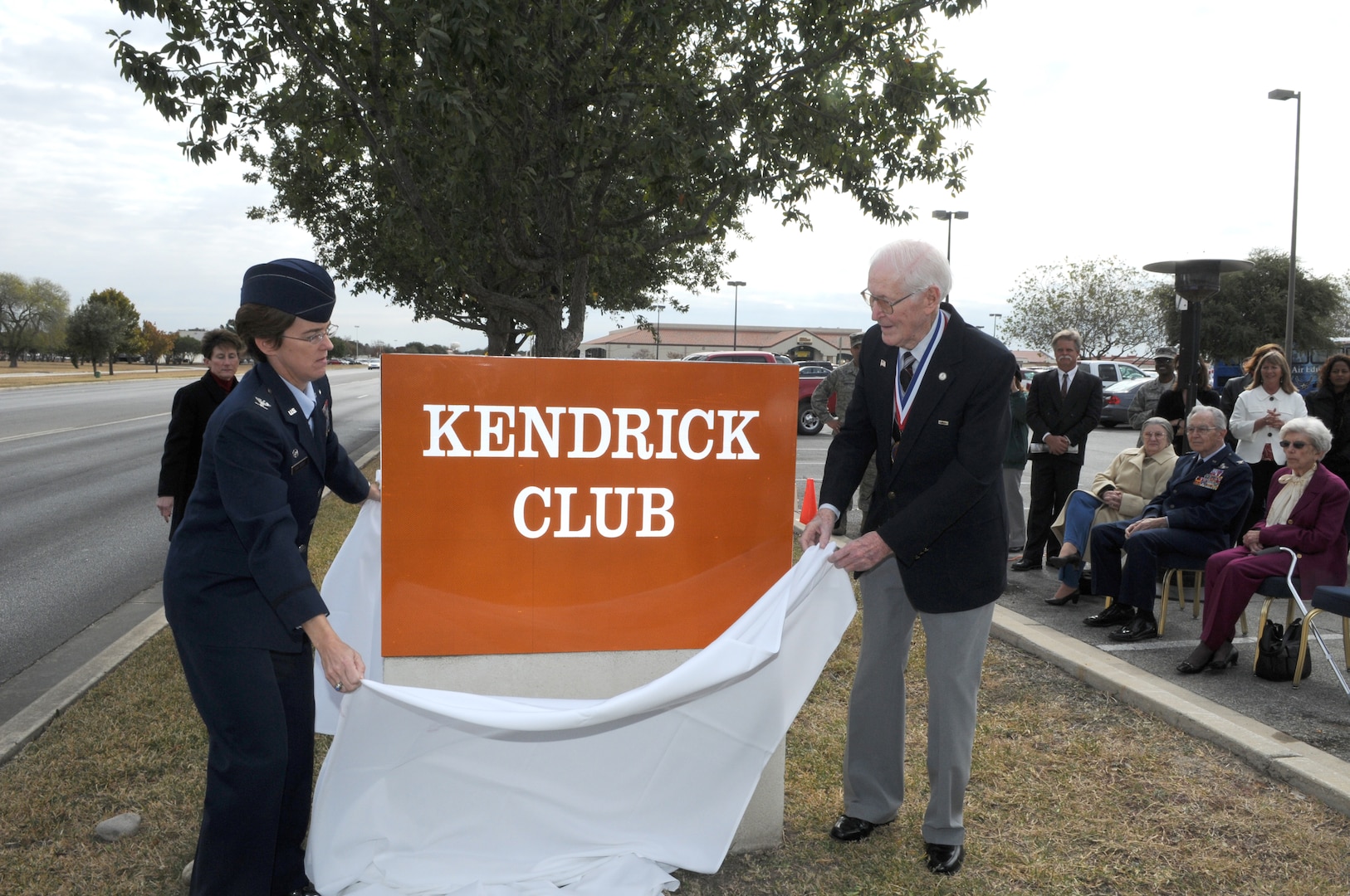 Col. Jacqueline Van Ovost, 12th Flying Training Wing commander, and retired Chief Master Sgt. Guy Kendrick lift the cover off the new enlisted club sign redesignating it the "Kendrick Club." (U.S. Air Force photo by Rich McFadden)