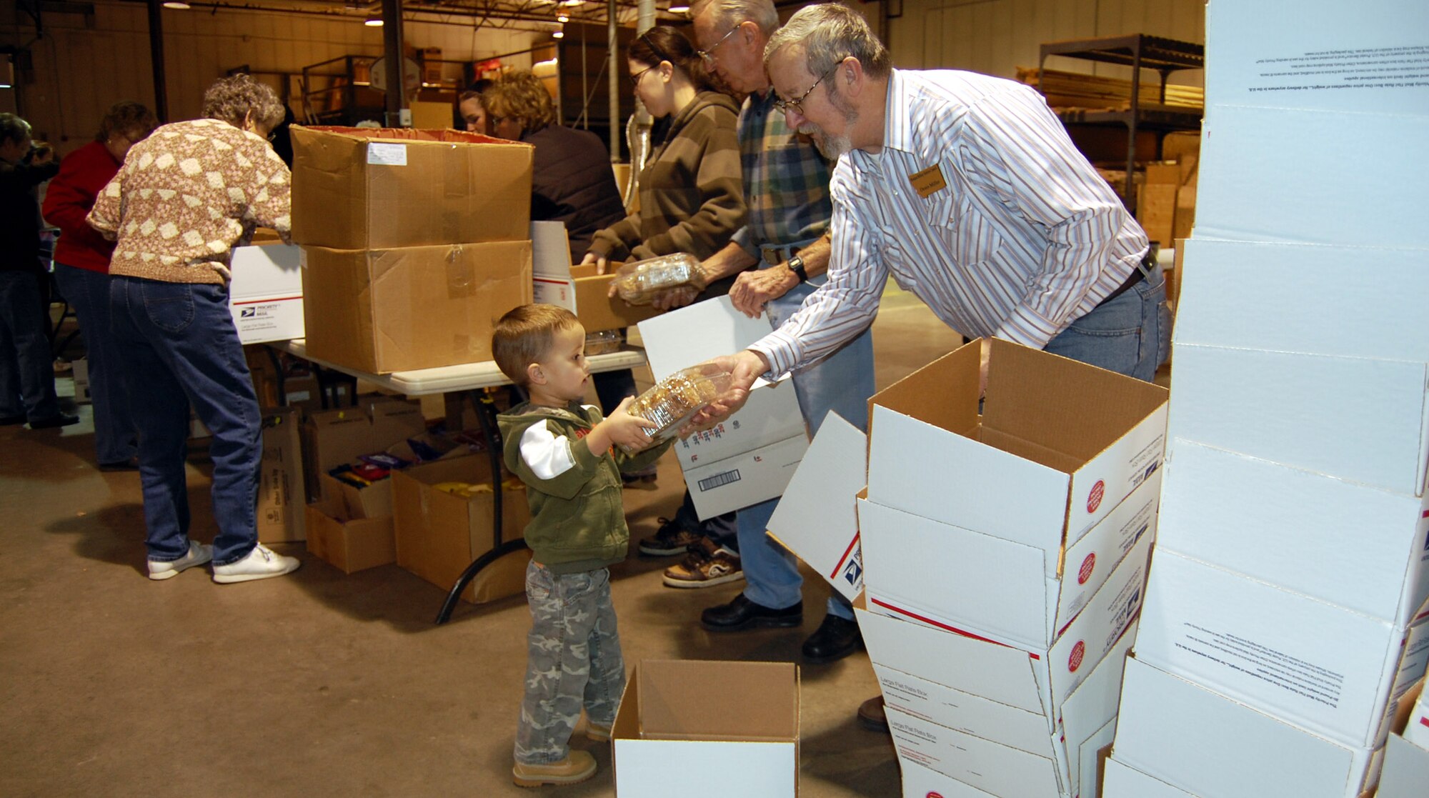 Little Warrior David, 3, hands Retiree Council member Denis Miller a package of homemade cookies Dec. 6 during the final phase of the 10th Annual Operation Happy Holidays. Volunteers and organizers packed more than 200 boxes and shipped them the same day. (U.S. Air Force photo/Valerie Mullett)