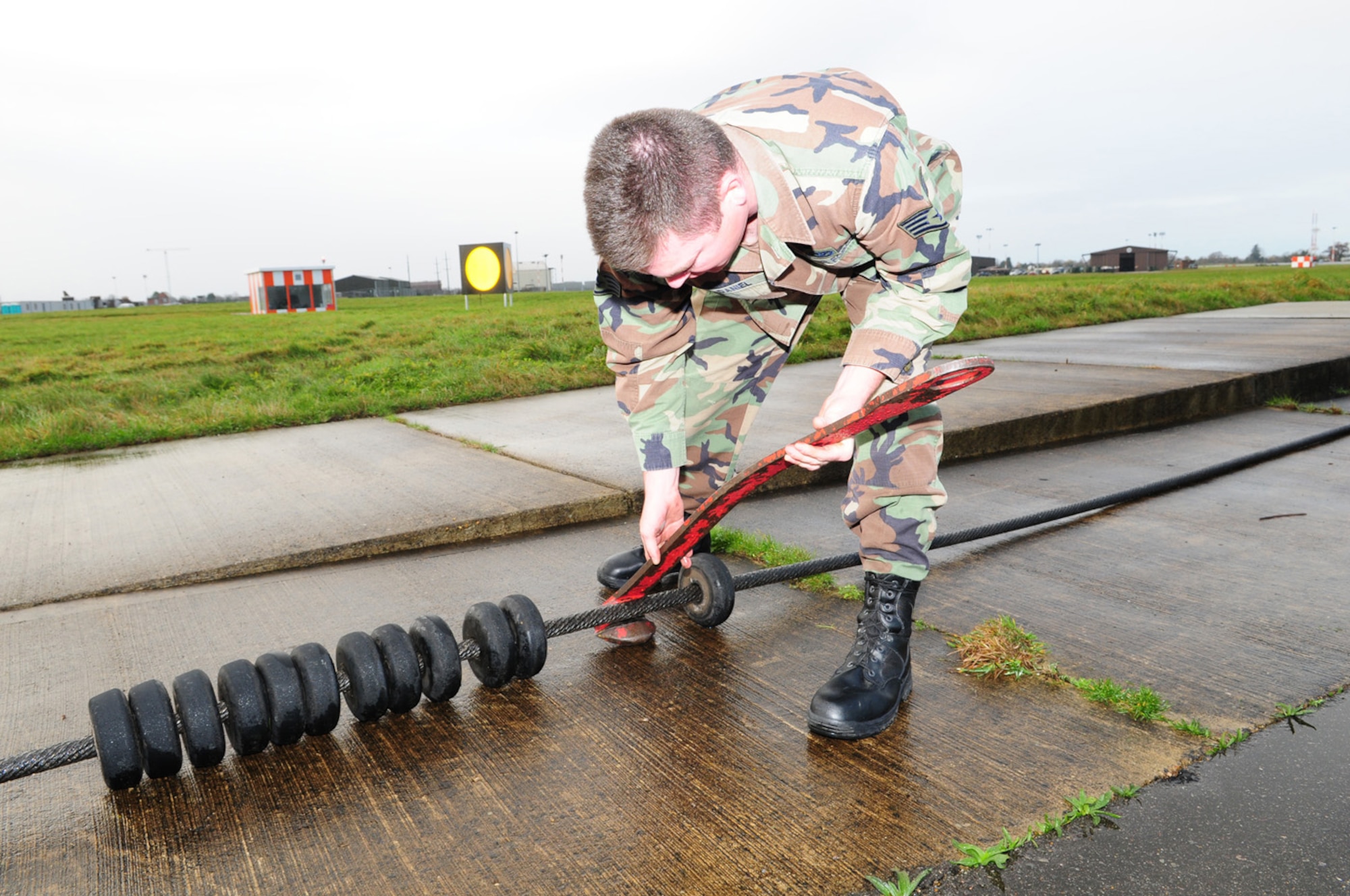 Staff Sgt. Ryan McDaniel, 100th CES Barrier Maintenance, fits a hook, similar to that on a fighter aircraft, onto a cable to demonstrate how the aircraft catches on and gets brought to a stop. The ‘donuts’ on the cable keep it 2 inches off the ground. The cables are checked each day to ensure they are not cracked or broken, have no kinks and are in good working order. If a call comes in to the barrier maintenance personnel, they can be at the barrier shacks and have everything in place and cables set up in about seven minutes. Sergeant McDaniel they said their goal is a three minute response time from Building 705, on the south side of base, to any of the barrier shacks on base. While there have been no actual barrier engagements in the past year, there were two certification engagements – with two aircraft from RAF Lakenheath (one at each end of the runway) – in September. (U.S. Air Force photo by Karen Abeyasekere)