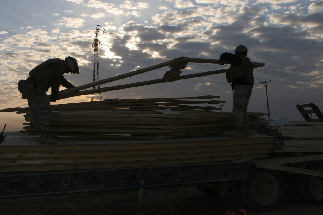 Two Marines with General Support Platoon, Company B, 1st Combat Engineer Battalion, Regimental Combat Team 5, make use of the early sunrise light and unload pre-fabricated rafter forms from the back of a trailer near the city of Haditha, Iraq, Dec 15. The engineers worked in three teams of cutting, off-loading and constructing in order to erect the wooden huts in a timely fashion. The huts will be used by Iraqi Security Forces who will man a nearby Traffic Control Point. ::r::::n::