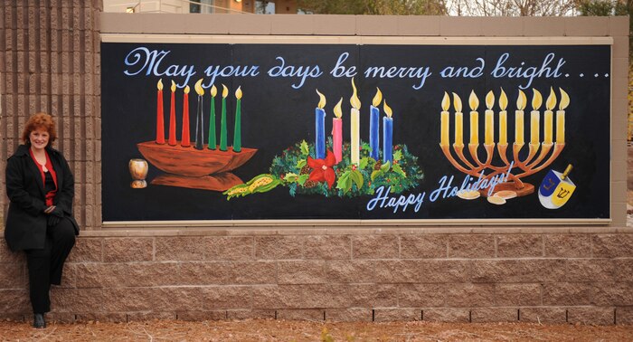 Mary Jo Wheeler, 99th Communications Squadron telecommunications project manager at Creech AFB, poses with the sign she painted at the Nellis Air Force Base, Nev., front gate on Dec. 17, 2008. The holiday-themed sign shows five advent, nine menorah and seven kwanza candles. (U.S. Air Force Photo / Airman 1st Class Stephanie Rubi)