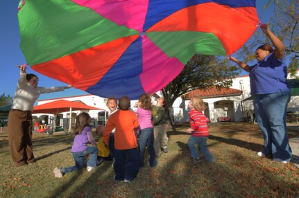 Child Development Center staff members Theresa Duncan, left, and Allison Dixon hold a multicolored parachute aloft recently, as children play underneath. (photo by Mellissa Peterson)