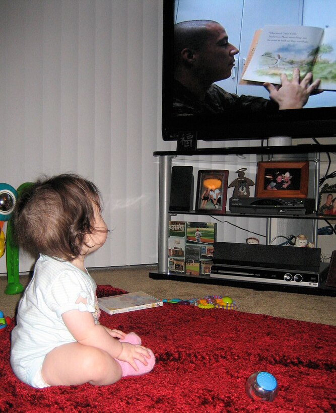 Myah Ayn Cano looks on as her father reads a book to her while he was deployed.  Service members can make recordings of reading books to children while they are deployed through United Through Reading.  The program allows service members to stay apart of their children’s lives when they are across the world.::r::::n::::r::::n::