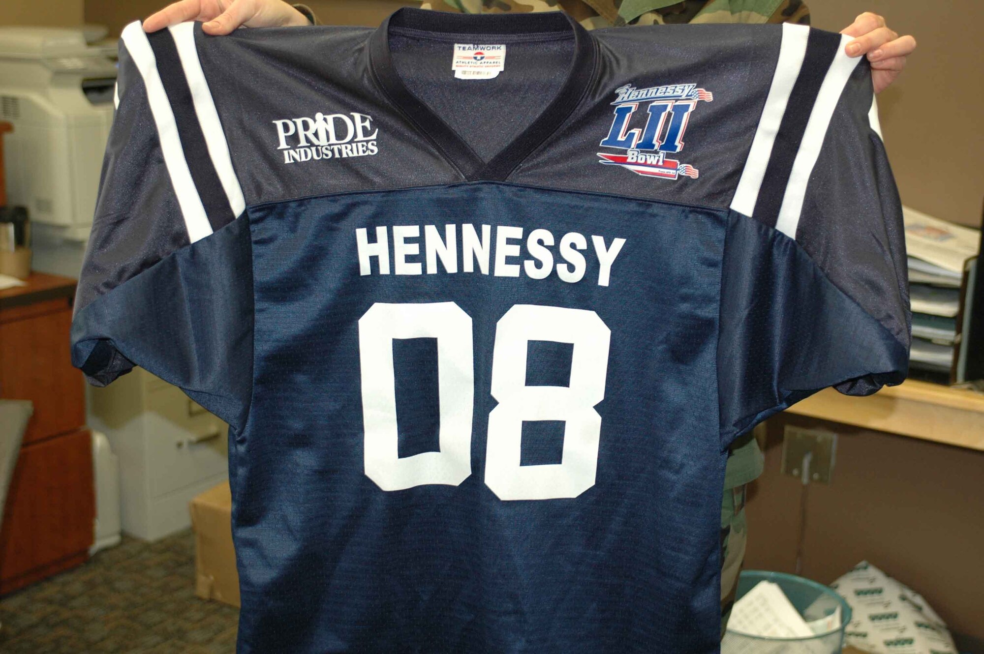 Staff Sgt. Ginger Warner, noncommissioned officer in charge of the Sierra Inn Dining Facility’s storeroom, holds up a Hennessy Bowl jersey. The Hennessy Bowl concept was concocted by dining facilities leadership as a means to motivate its members to capture the Hennessy Award, an honor bestowed upon the Air Force’s best dining facility. (U.S. Air Force photo/Nick DeCicco)