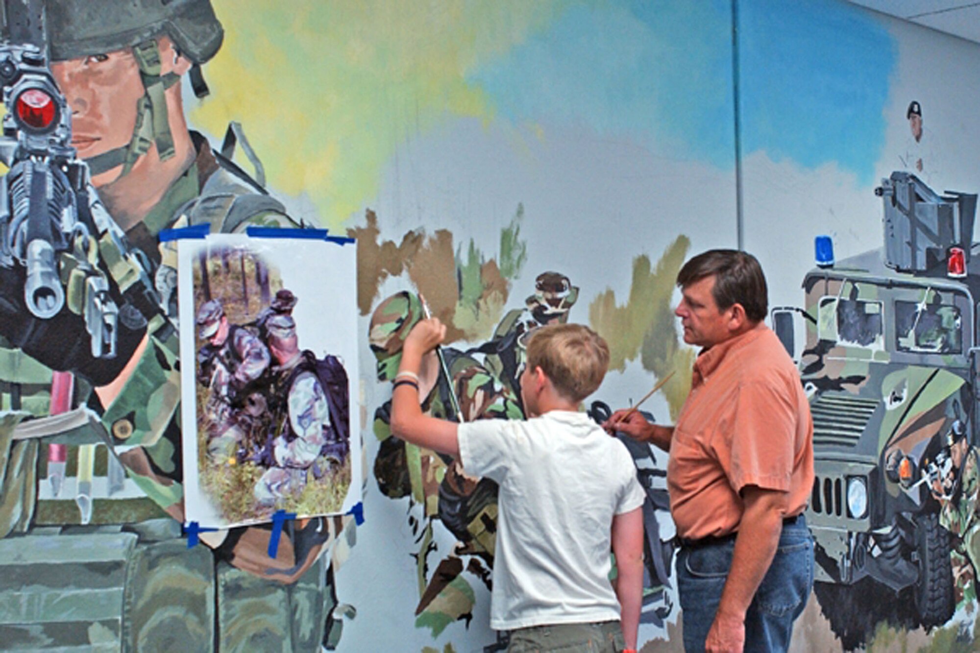 Bob Stillwell, 341st Communications Squadron contracted graphic artist, gets help from his grandson Logan, while painting a mural for the 341st Security Forces Group headquarters. Mr. Stillwell volunteered more than 230 hours of his off-duty time to paint the mural in honor of the security forces career field. His son Bobby flew in from Washington to help paint the background and HUMVEE, while Logan, who is an aspiring artist, traveled down to Malmstrom from Conrad, Mont. (U.S. Air Force photo)