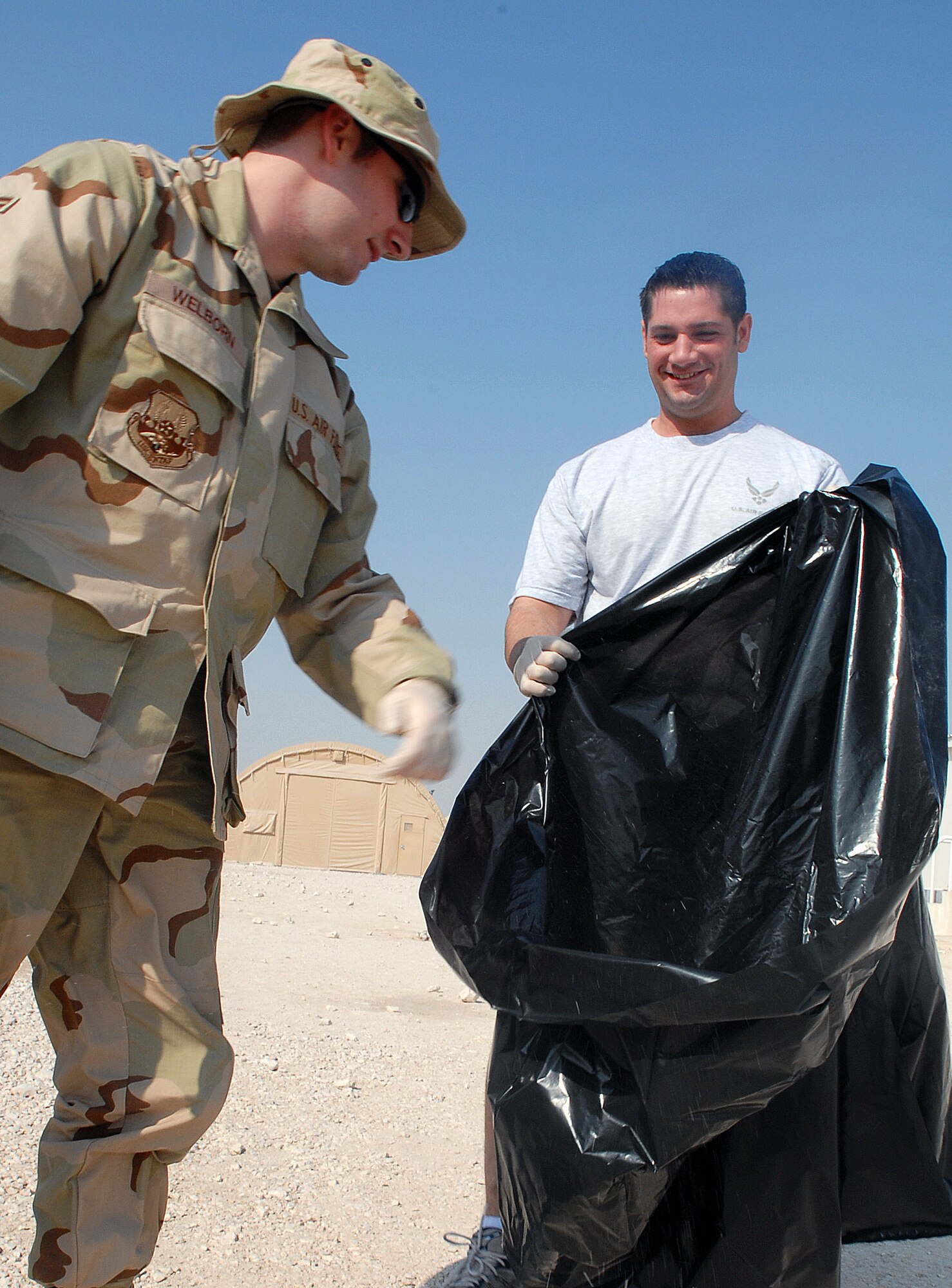 SOUTHWEST ASIA-- Airman 1st Class Jeremi Welborn places garbage into a trashbag Staff Sgt. Joshuah Jacobs is holding at a Southwest Asia air base Jan. 20. Both Airman participated in the Desert 5's first Coalition Compound Cleanup, an event in which volunteers helped improve the appearance of the area around transient tents for incoming occupants. The Desert 5 works with the Top 4, the First Sergeants, and Wing leadership to provide Airmen a forum to discuss issues that affect the morale of base members. In addition, the Desert 5 is actively involved in promoting esprit de corps through programs like the monthly Wing Promotion Ceremony and Mentorship Award program. (U.S. Air Force photo by Senior Airman Domonique Simmons) 