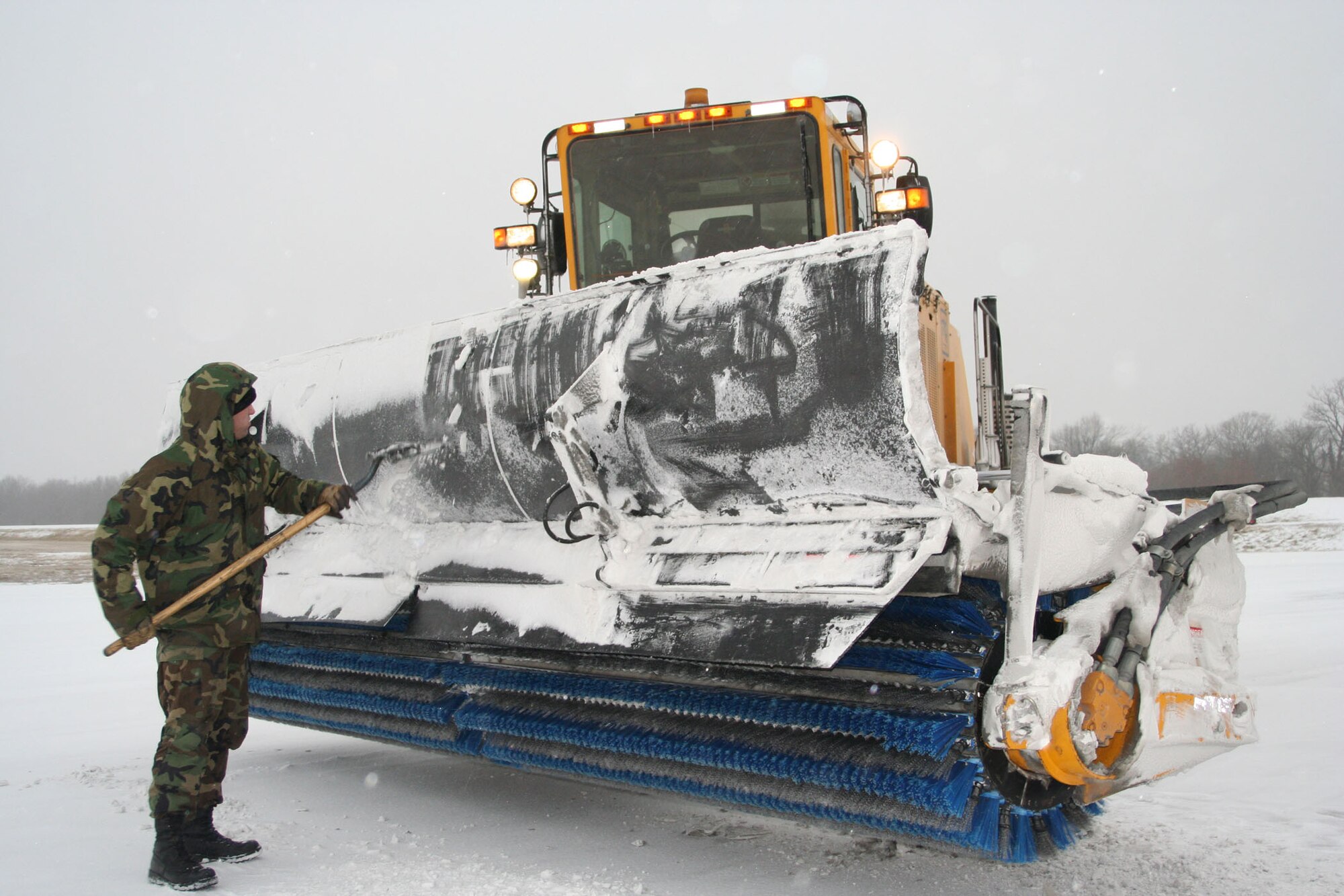 WHITEMAN AIR FORCE BASE, Mo. – Staff Sgt. Kevin Steinke, 509th Civil Engineer Squadron, scrapes snow and ice form the guard of a brush truck to prevent the rotating brush from jamming. The 509 CES snow removal crew applies a special chemical to the surface of the taxiway and runway prior to expected snow fall to ease removal process. (U.S. Air Force photo/Maj. Joe DellaVedova)