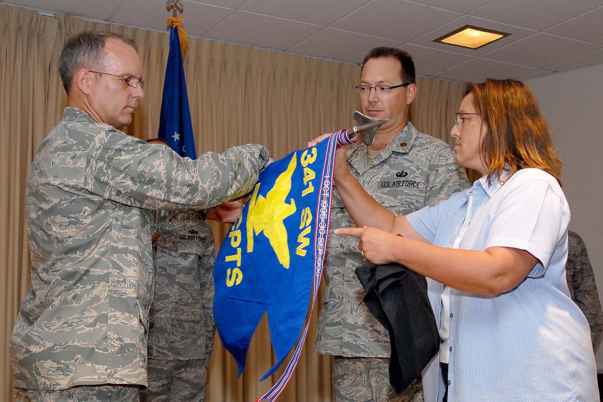 Ramona Rogers, financial analyst, and Col. Michael Fortney, 341st Missile Wing commander,  roll up the 341st Comptroller Squadron guidon before retiring it during a ceremony to merge their squadron with the 341st Contracting Squadron July 11. Maj. Trevor Whitehill, center, 341st CPTS commander will continue in that role for the combined 341st Comptroller Contracting Squadron. (U.S. Air Force photo/John Turner)