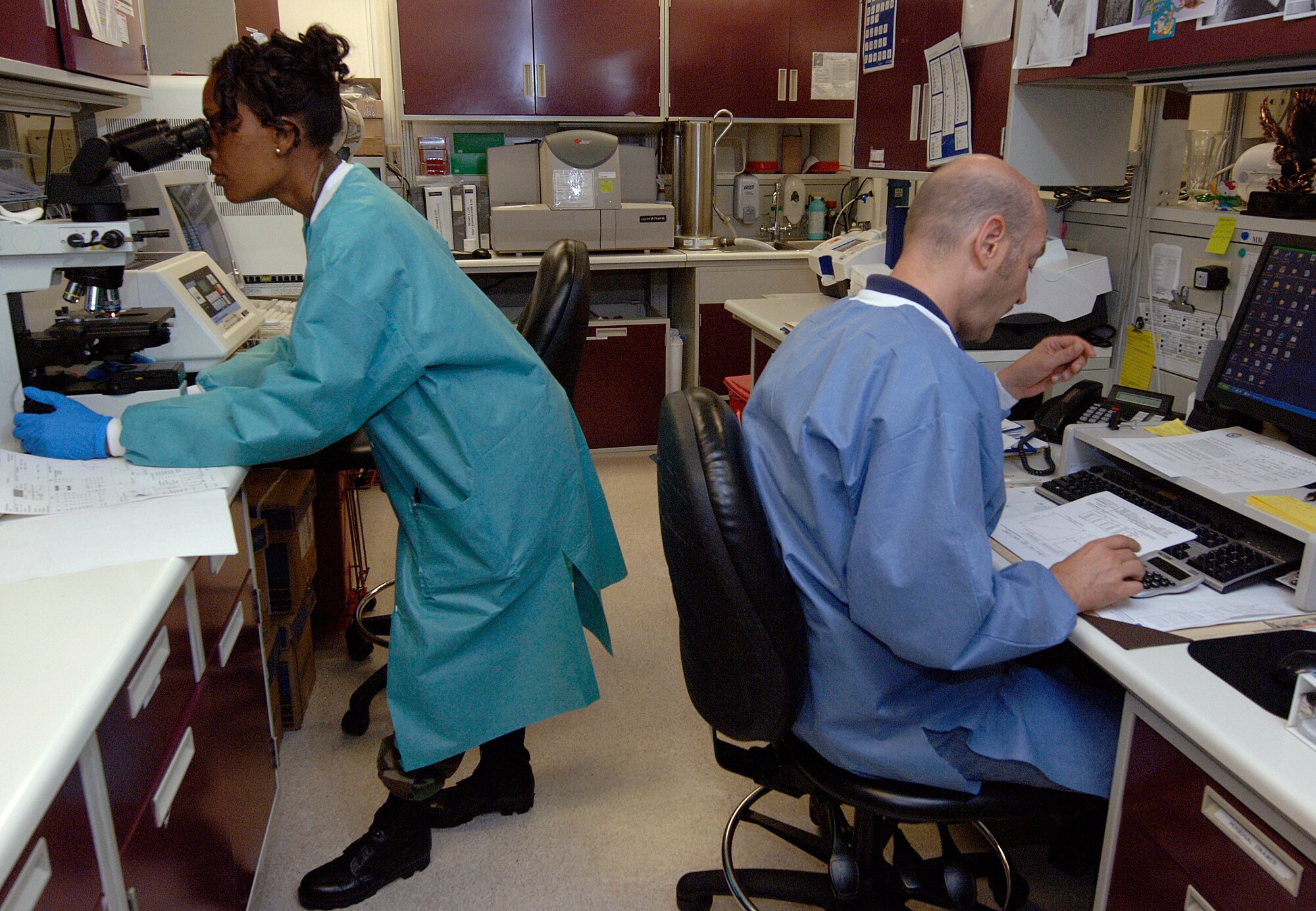 LANGLEY AIR FORCE BASE, Va. -- Tech. Sgt. Laronda Harris, the non-commissioned officer in charge of Hematology at 1st Fighter Wing Hospital, looks at specimens through a microscope in the microbiology section of the laboratory while Keith Johnson, chemistry technical supervisor, reads over laboratory test results July 15.  The microbiology section of the laboratory is where all cultures on various bodily samples are performed.  (U.S. Air Force photo/Airman 1st Class Sylvia Olson)