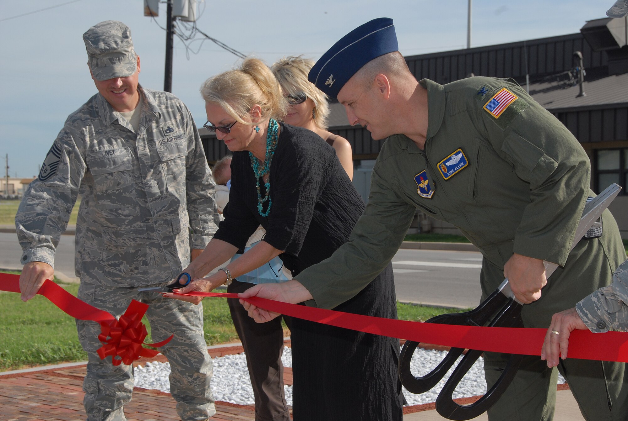 Chief Master Sgt. Kenneth Ivey (left) and Col. David w. Allvin, 97th Air Mobility Wing commander (right) assist Brenda Petzold in the ribbon cutting to open the Altus Air Force Base Airman Leadership School Brick Layer that will honor enlisted heroes and individuals that significantly impacted the lives of Airmen. (U.S. Air Force/A1C Cherice Bryant)