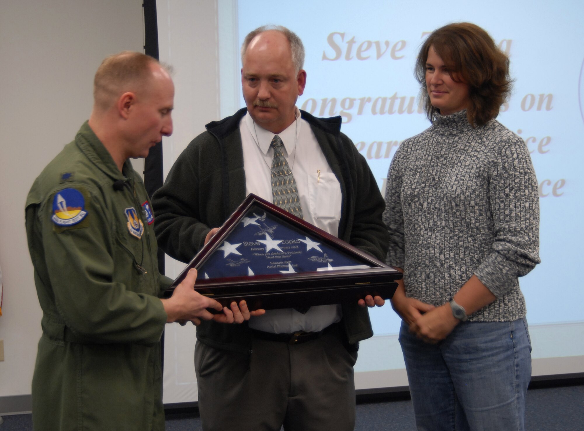 Lt. Col. Carl Schaefer, 445th Flight Test Squadron commander, presents a memento to Steve Zapka, Rohmann Services Inc. project manager, as Bobbi Zapka, aerial photographer and Mr. Zapka's wife, looks on during his 25-year celebration at Edwards. Mr. Zapka has more than 5,000 flying hours as an aerial photographer. (Air Force photo by Airman 1st Class Stacy Sanchez)