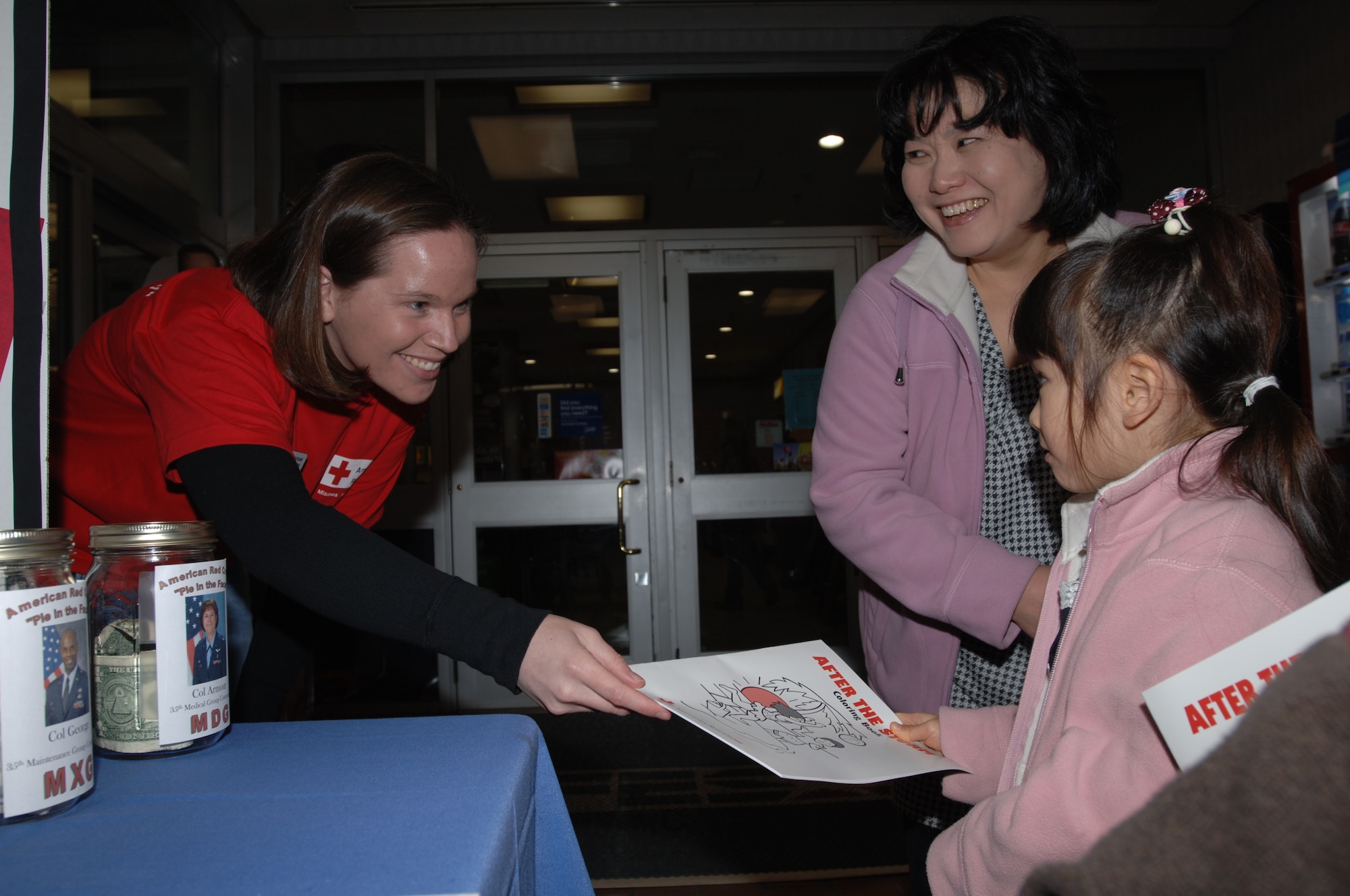 MISAWA AIR BASE, Japan -- Mio Watamabe, 4 years old, accepts a Red Cross coloring book from Maggie Kuntz, Red Cross chairman of volunteers, while her mother Hiroko Watamabe watches at the Red Cross information booth near the commissary March 20, 2008.  The Red Cross is holding several classes on first aid, babysitter training, and volunteer orientation this month.  (U.S. Air Force photo by Senior Airman Robert Barnett)