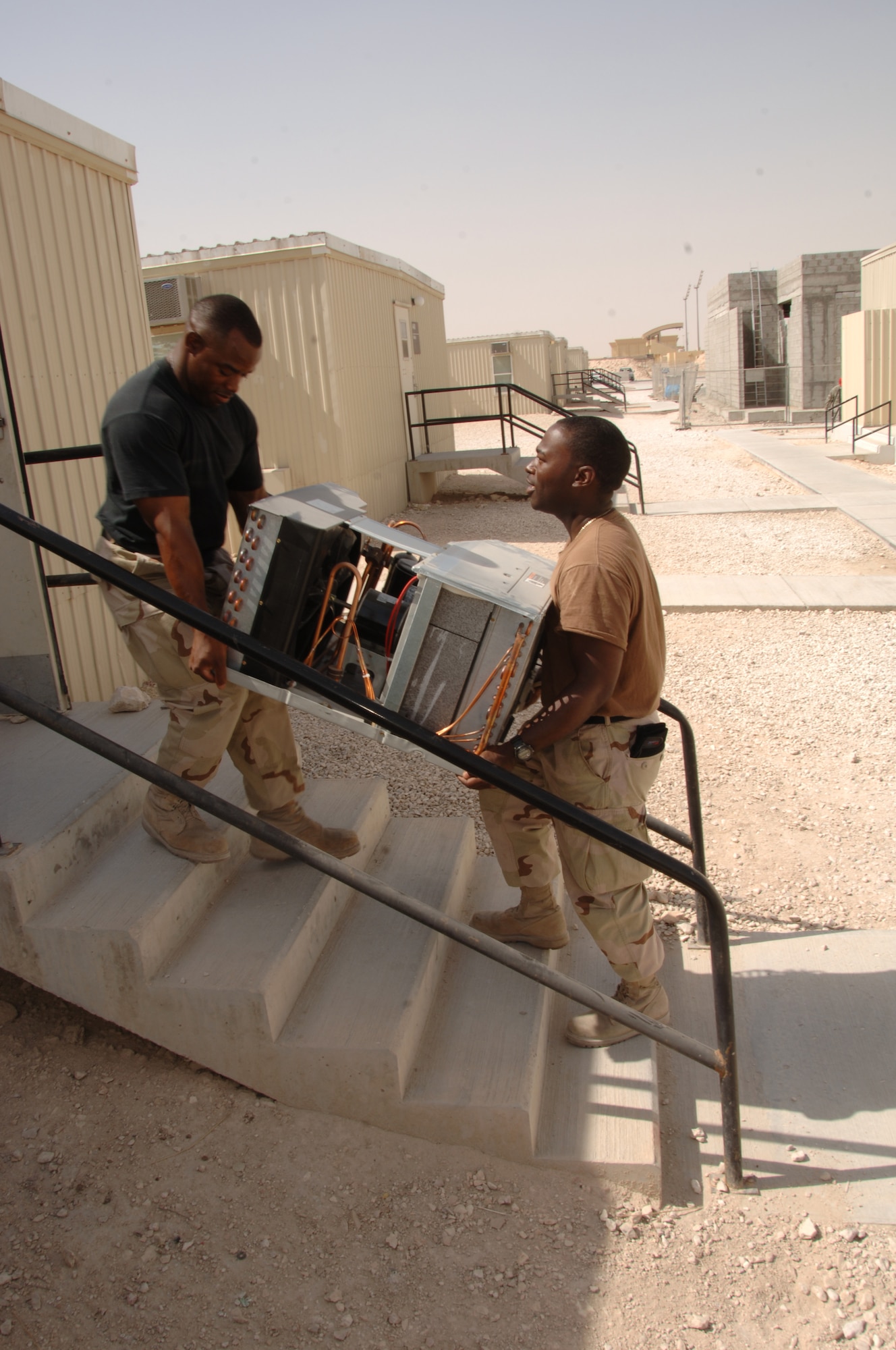 Staff Sgts. Dwayne Carter (left) and Andrew Baines, 379th Expeditionary Civil Engineer Squadron heating, ventilation and air conditioning technicians, carry a new air conditioning unit to be installed in a dormitory. Sergeant Carter, deployed from Ramstein Air Base, Germany, and Sergeant Baines, deployed from Andrews Air Force Base, Md., perform the semiannual preventive maintenance procedure on window air conditioning units throughout coalition compound to prevent dorm fires. (U.S. Air Force photo/Senior Airman Tong Duong)