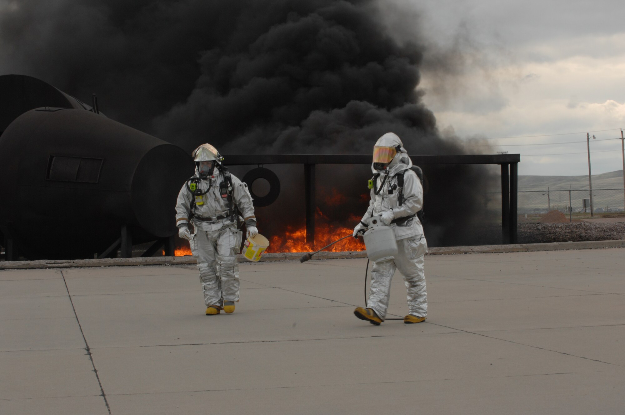 (Left) Staff Sgt. Raymond Granillo, 28th Civil Engineer Squadron fire protection crew chief, holds a bucket of flammable liquid used to ignite training fires while Staff Sgt. Chris Voth, 114th Air National Guard Civil Engineer Squadron fire protection crew chief, turns his propane tank off here, May 13.The pit fire exercise helps satisfy certification requirements for firecrews. (U.S. Air Force photo/Airman 1st Class Adam Grant) 
      
