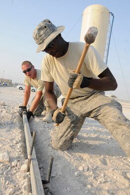 Airman 1st Class Matthew Lawrence, heavy equipment operator assigned to the 379th Expeditionary Civil Engineer Squadron, positions a beam while Airman 1st Class Antonio Fox, heavy equipment operator assigned to the 379th Expeditionary Civil Engineer Squadron, uses a sledgehammer to drive the stake into the ground so they can form up an area and pour concrete to create a walking path Nov. 13, at an undisclosed air base in Southwest Asia. Airman Lawrence, a native of Richmond, Va., is deployed from Grand Forks Air Force Base, N.D. and Airman Fox, a native of Salisbury, N.C., is deployed from McConnell AFB, Kan. Both are deployed in support of Operations Iraqi and Enduring Freedom and Joint Task Force-Horn of Africa. (U.S. Air Force photo by Staff Sgt. Darnell T. Cannady/Released)