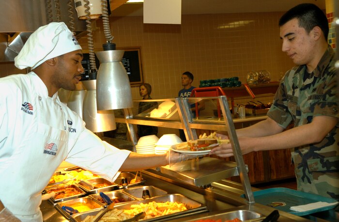 Senior Airman John Jackson passes a hot plate to Airman 1st Class James Macias of the 57th Component Maintenance Squadron during lunch rush at the Crosswinds Dining Facility here Nov. 18. The 99th SVS won the Air Combat Command Hennessy award and is now competing on the Air Force level. (Air Force photo by Senior Airman Oleksandra Manko)