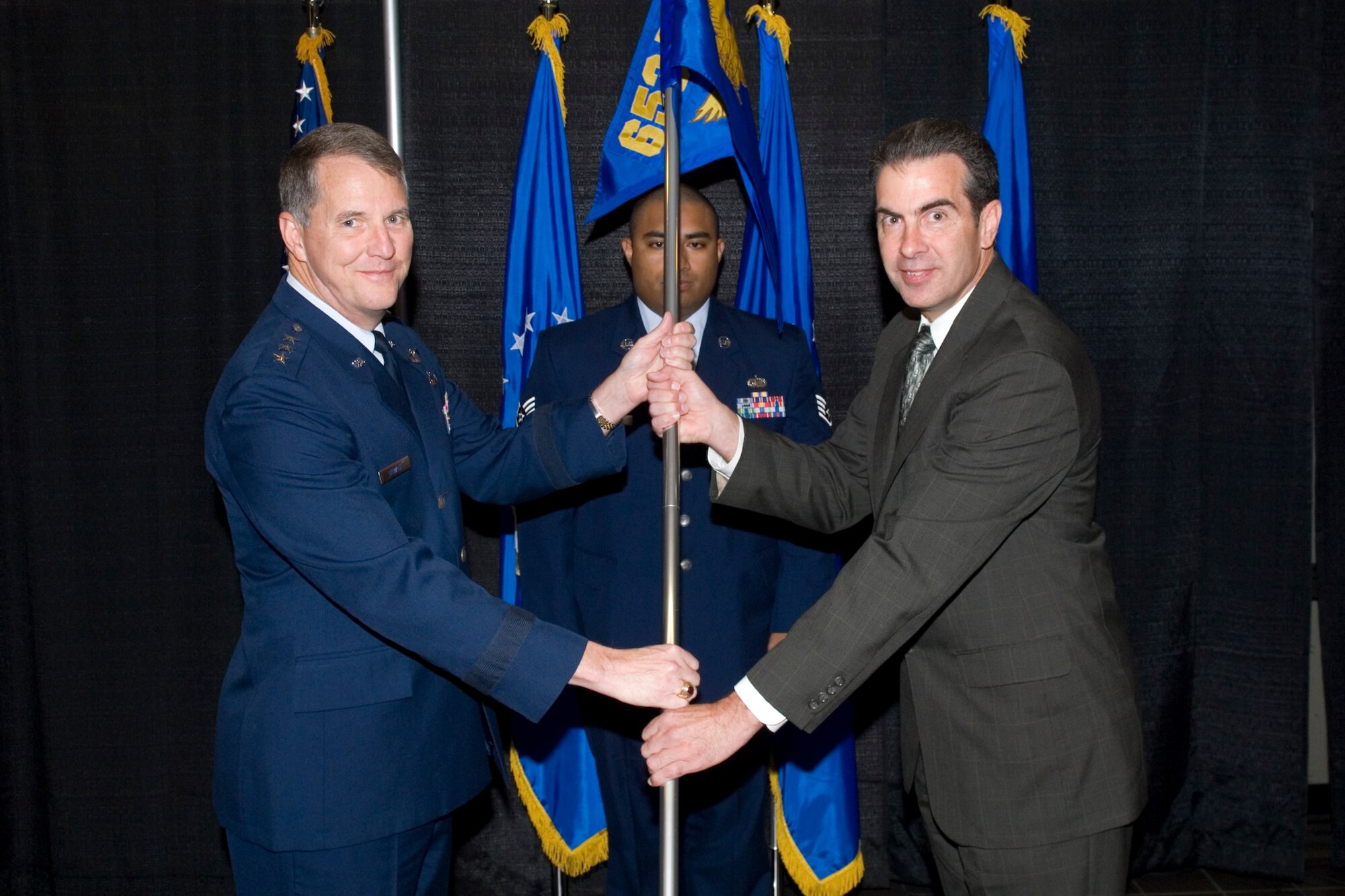 Electronic Systems Center Commander Lt. Gen. Ted Bowlds passes the 653rd Electronic Systems Wing guidon to the wing’s incoming director, Senior Executive Service member Ron Mason, during an Assumption of Leadership ceremony Oct. 2, as Staff Sgt. Patrick Raass looks on. Mr. Mason, during his remarks, acknowledged the great successes that have been achieved by the 653 ELSW and pledged to work tirelessly to support the wing’s mission and people. (Photo by Rick Berry)
