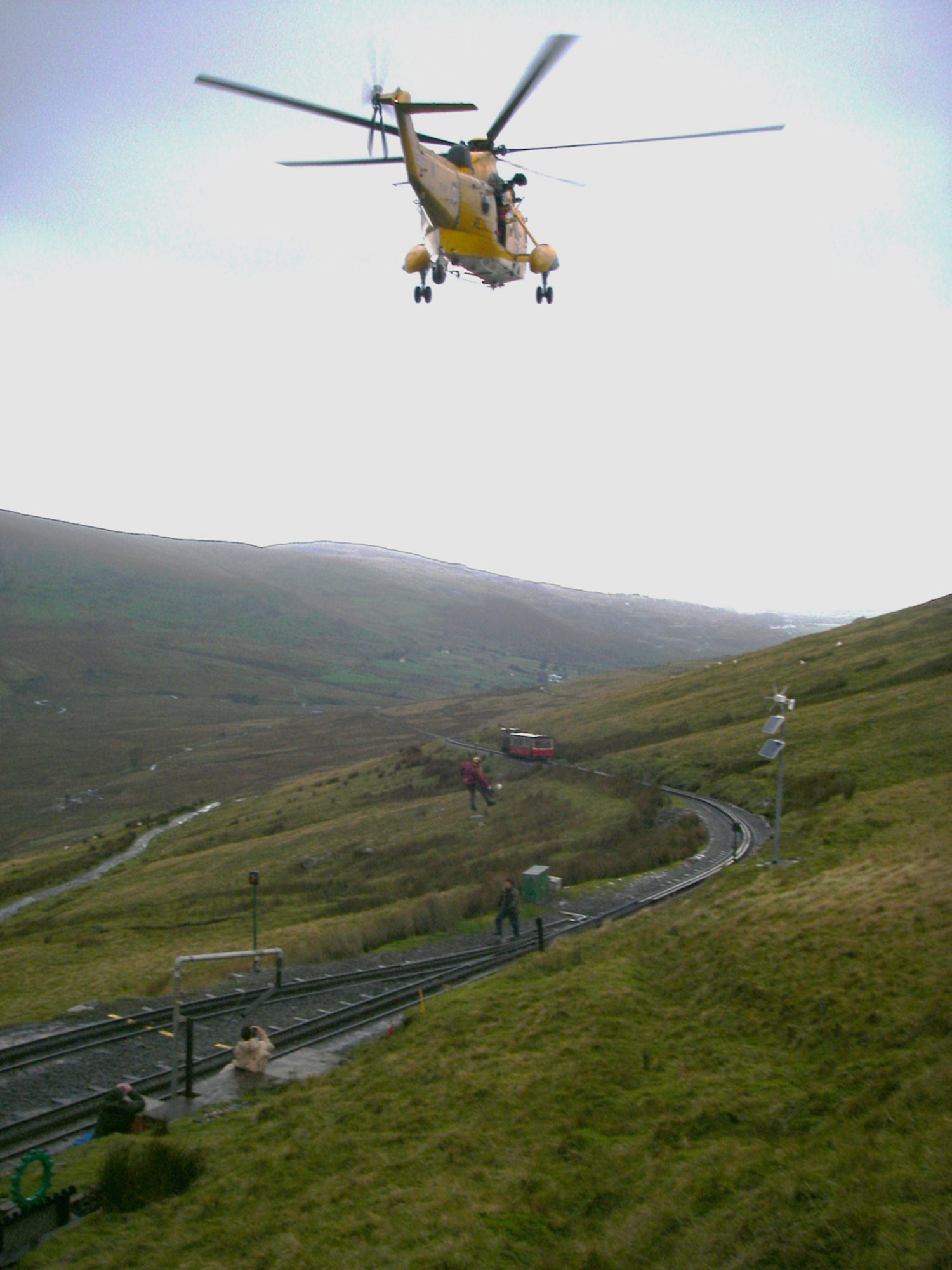 A rescue helicopter from 22 Squadron, RAF Valley, Wales, winches up the casualty from halfway down Mount Snowdon, ready to take him to the hospital. Three Defence Fire Service firefighters, Alan Coldwell, Chris Gould, and Jerry Miles, all 100th Civil Engineer Squadron Fire Department, and their mountain guide, Dave Wainwright, 100th Force Support Squadron Outdoor Recreation, rescued the man they found alone and suffering from hypothermia and frostbite at the peak of Mount Snowdon Oct. 3. After calling emergency services, the four helped bring him halfway down the mountain, with the help of passengers on a train from Snowdon Mountain Railway. The train never usually runs at this time of the year as the weather is too bad, but by coincidence, it was taking passengers to the top of Mount Snowdon at the same time the firefighters found the man. (Courtesy photo by Doug Blair, Snowdon Mountain Railway)