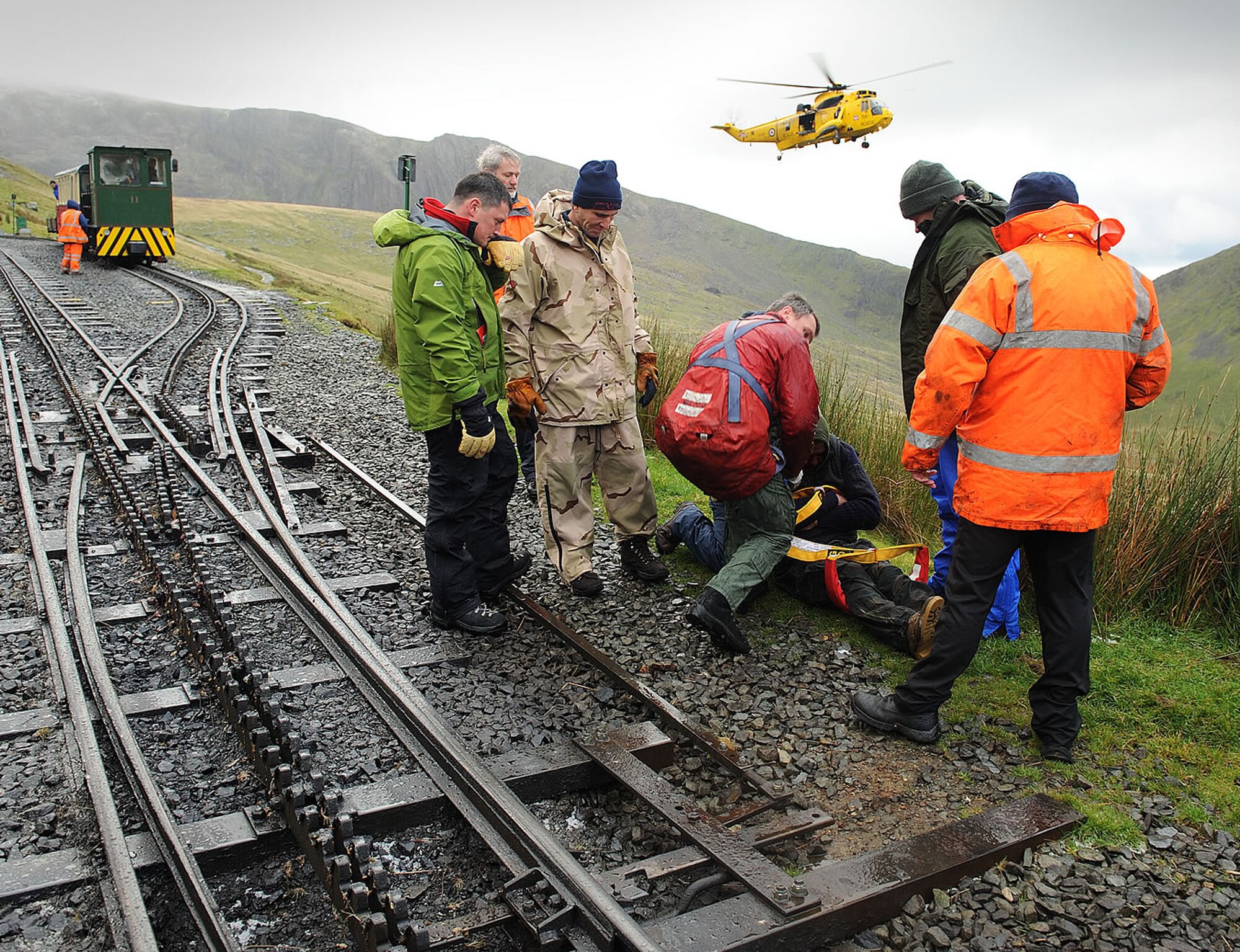 Dave Wainwright, 100th Force Support Squadron Outdoor Recreation director, and Defence Fire Service firefighters Chris Gould, Jerry Myles and Alan Coldwell, all 100th Civil Engineer Squadron, look on with members of the Snowdon Mountain Railway to ensure the casualty they rescued from the top of Mount Snowdon is safely prepared by the winchman to be taken up into the rescue helicopter from 22 Squadron, RAF Valley, Wales.(Courtesy photo from Snowdon Mountain Railway)