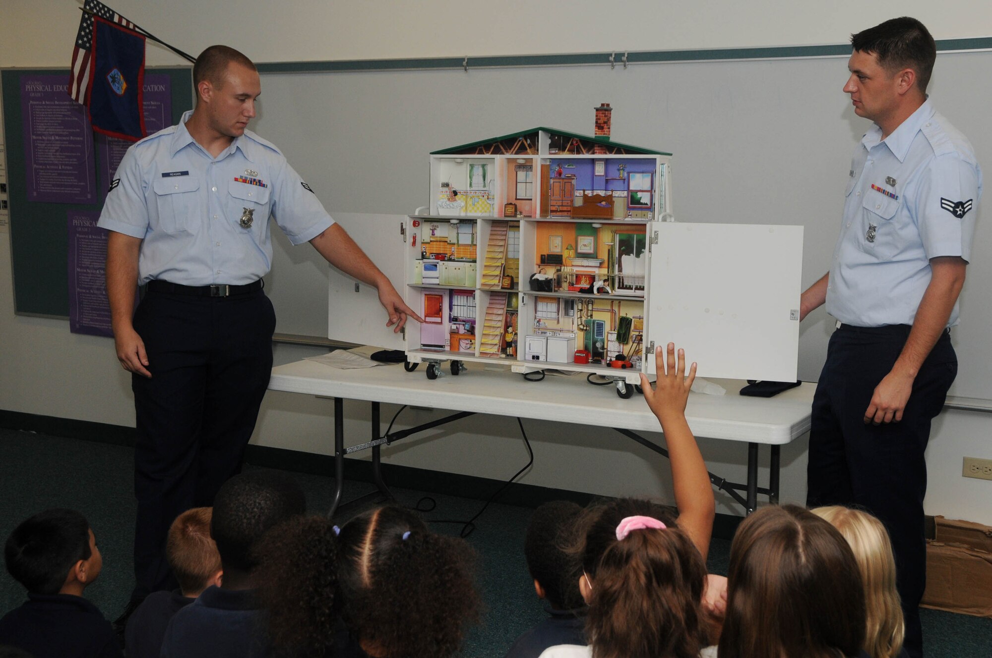 ANDERSEN AIR FORCE BASE, Guam - Airmen 1st Class Sean Reagan and Geron Ramsdell, 36th Civil Engineer Squadron firefighters, demonstrate how to respond to several fire mishaps within the home during Fire Prevention Week here Oct. 9. The dollhouse used for this exercise produces simulated smoke and lights representing fire to better engage with the students. (U.S. Air Force photo by Senior Airman Sonya Croston)