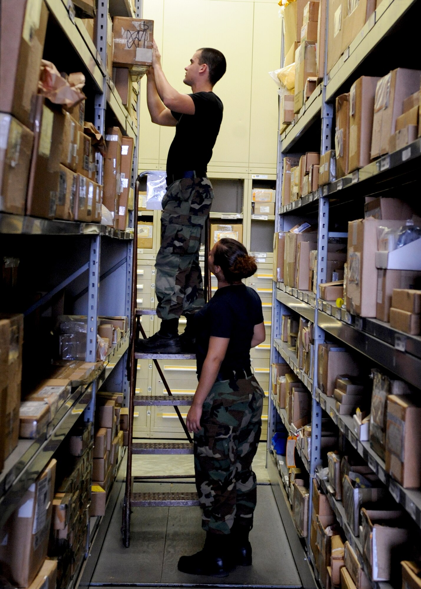 Airman 1st Class John Peppers, member of the Aircraft Parts Store (APS), places aircraft material on shelves at Seymour Johnson Air Force Base, North Carolina on October 8, 2008.  Airman 1st Class Giselle Giron practices safety by standing at the bottom of the stairs incase A1C Peppers falls. (U.S. Air Force photo by Airman 1st Class Whitney Stanfield)