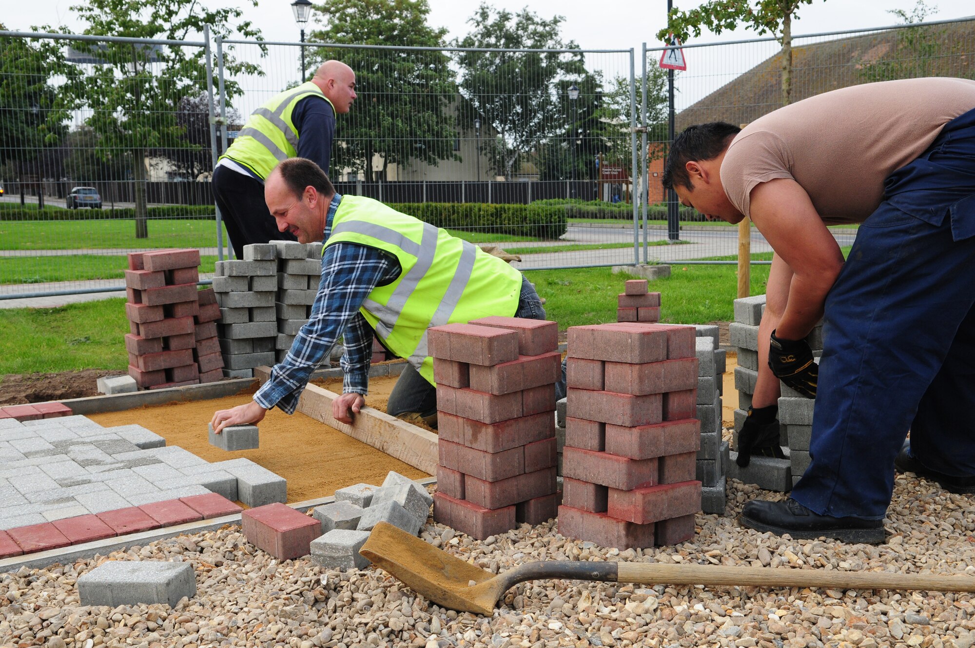 Richard Austin, center, lays bricks in the pathway of "Holiday Park" -- an area specifically for the annual holiday tree and cards display -- as Phil Winder and Airman Justin Cather, all 100th Civil Engineer Squadron, move bricks used in the construction. The area will have sidwalks in a hexagon shape, with five live spruce trees in the middle. The area should be finished by the end of October. (U.S. Air Force photo by Karen Abeyasekere)