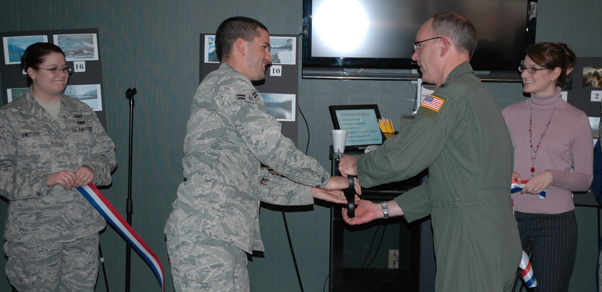 Airman 1st Class Anthony Melendez, 341st Missile Maintenance electro-mechanical team member, and Col. Michael Fortney, 341st Missile Wing commander, finish cutting a ribbon with a pair of over-sized scissors at the re-opening of the Detour Airmen's Center. Senior Airman Katelyn Switzer, 341st MW chaplain assistant and Julie Wiseman, Detour coordinator watch and hold the ribbon. (U.S. Air Force photo/Senior Airman Dillon White)