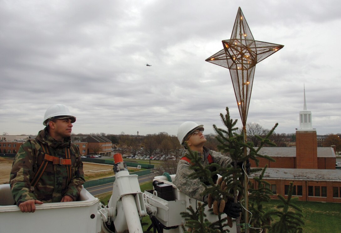 Airman 1st Class Victor Gonzalez Dias, 316th Civil Engineer Squadron electrical systems apprentice, left, observes as Airman Jonathan Smith, 316 CES electrical systems apprentice, right, straightens the star atop the holiday tree near Chapel I prior to the Christmas tree and Hanukkah Menorah Lighting Ceremony Wednesday. (U.S. Air Force photo/ Bobby Jones)