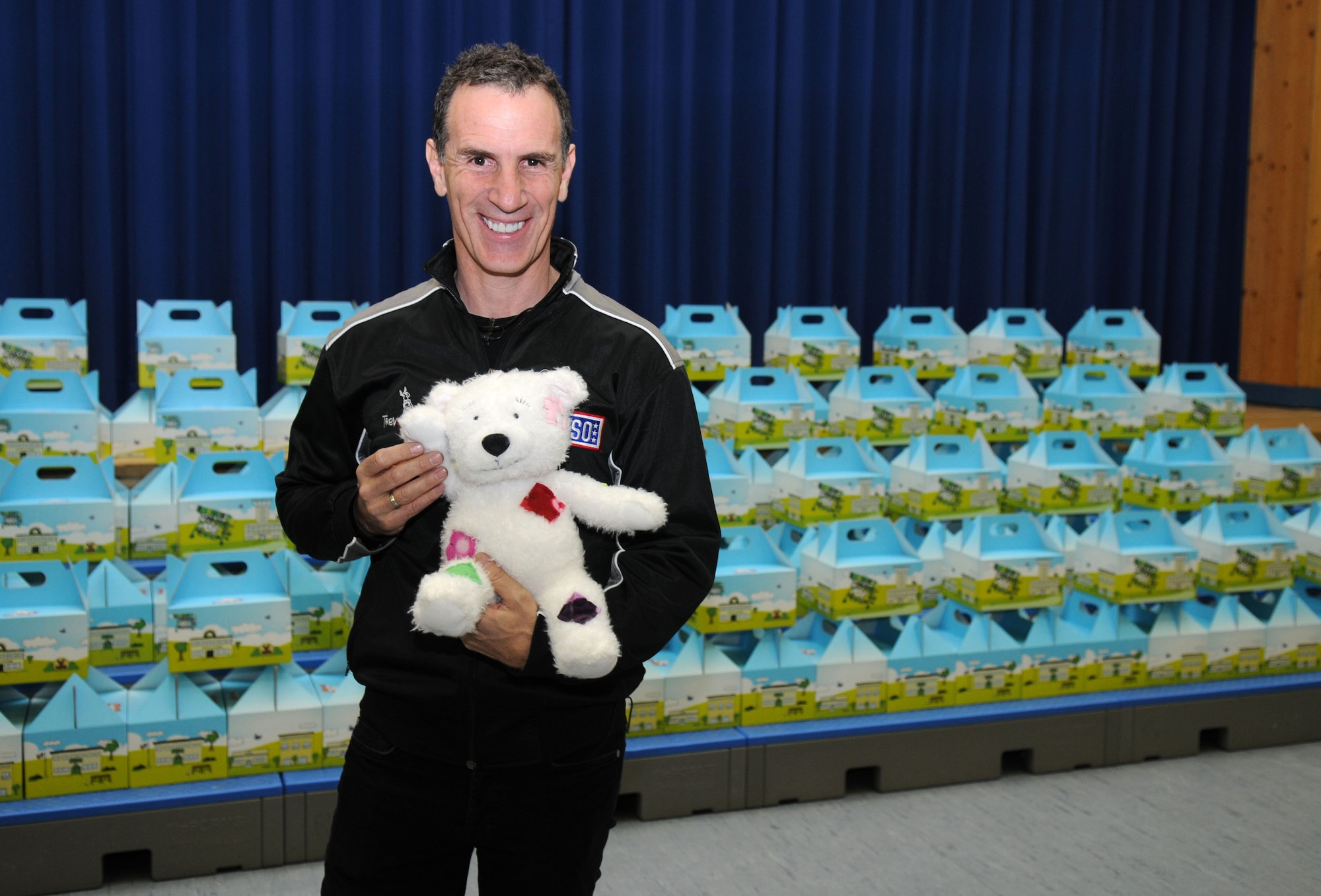 Mr. Trevor Romain, children's author, proudly displays his new character Cuzzie the Bear in front of Cuzzie Cares Deployment Kits at Ramstein Elementary School, Ramstein Air Base, Germany, Dec. 17, 2009. Cuzzie is an inventive bear that creates flying machines with the help of his ground crew and is the centerpiece of a new kit designed to comfort school-age children while their parents are deployed. (U.S. Air Force photo by Airman 1st Class Brittany Perry)
