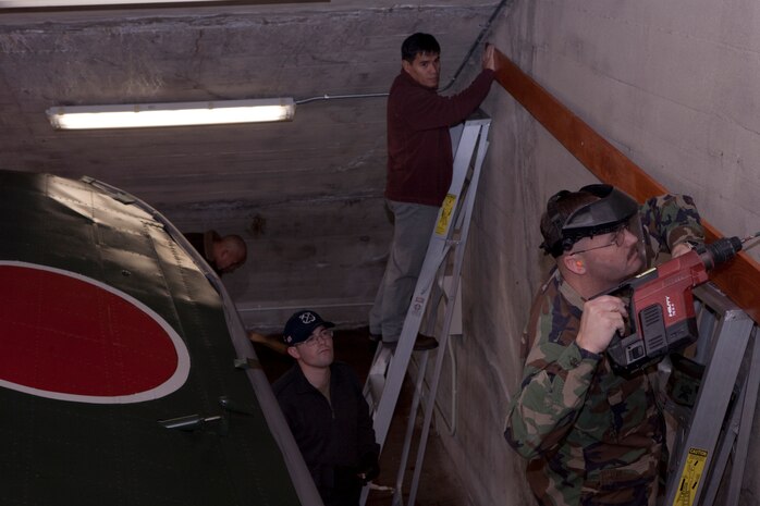 Petty Officer 1st Class Bobby Wilson drills a hole into the wall of Zero Hangar to mount a board for holding pictures during a Zero Hangar cleanup and beautification here Dec. 17. By hanging pictures on the walls, the volunteers hoped to free up walking space for patrons visiting the Zero Hangar display.