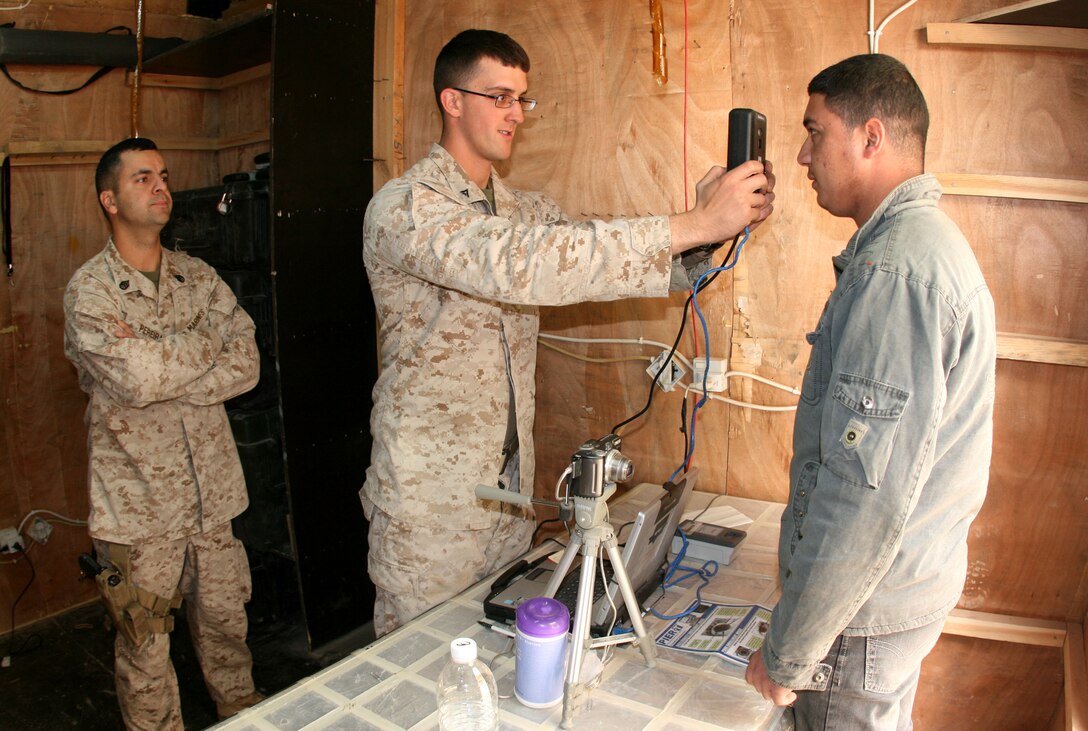 Staff Sergeant Rock Pereira (left), the staff non-commissioned officer in charge of processing badges and identification for the camp commandant’s office at Camp Korean Village, Iraq, supervises as Lance Cpl. Joseph Davieau records biometrics data for an Iraqi worker aboard the base Feb. 10.  Pereira, 31, of Queens, N.Y., is a detective with the New York City Police Department in his civilian career. Davieau, 23, is a full-time university student from Boonton, N.J. (Official USMC photo by Capt. Paul L. Greenberg)