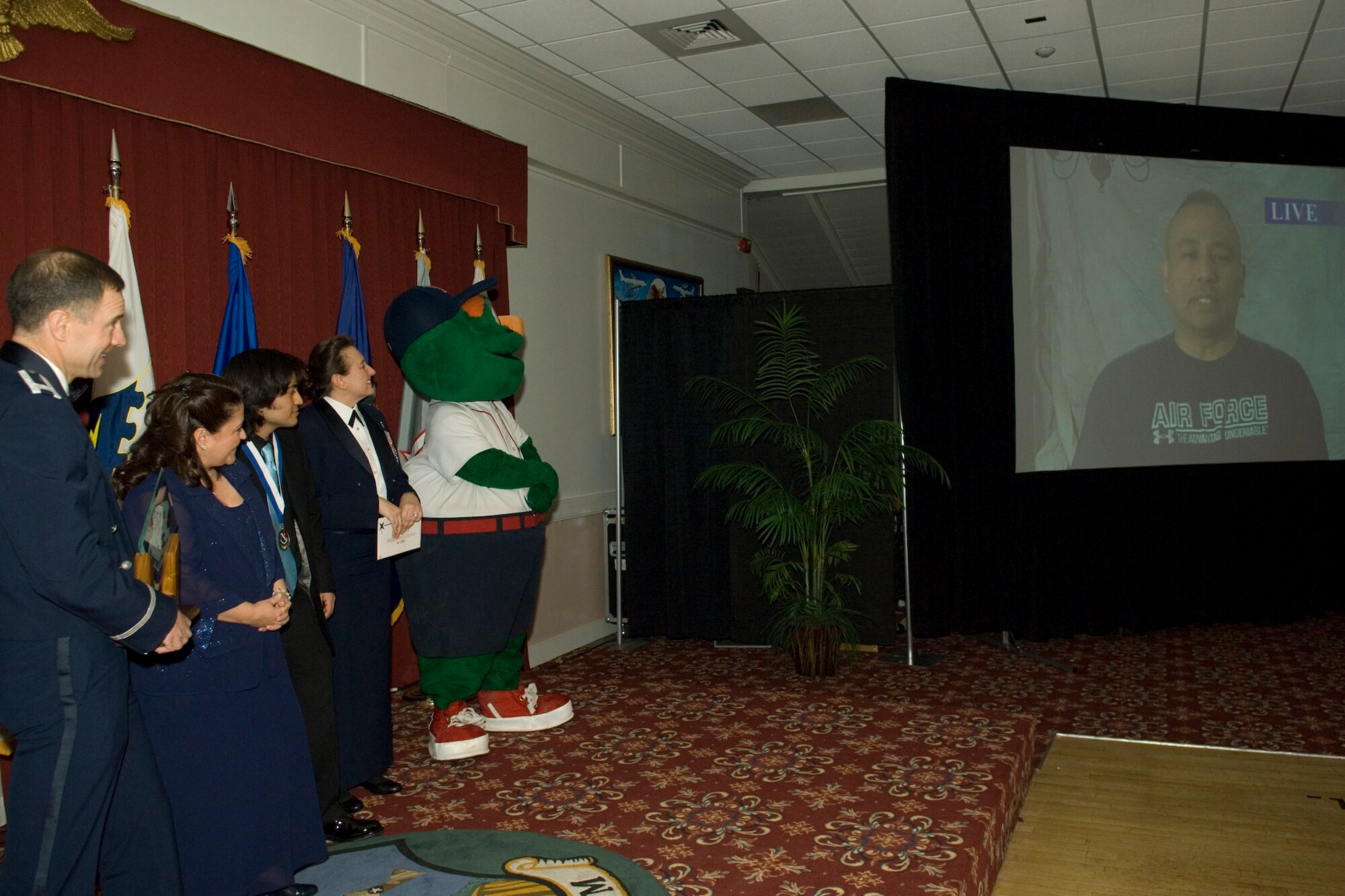 HANSCOM AIR FORCE BASE, Mass. – Senior Master Sergeant Raul Ruiz, 66th Mission Support Squadron first sergeant, speaks by video to the crowd gathered for the 66th Air Base Wing Annual Awards banquet Feb. 6, looking on are from left to right, Colonel Orr, 66th Air Base Wing commander, Sergeant Ruiz’s wife Brandi and son Ryan, Chief Master Sgt. Karen McCoy, ESC Command Chief and Boston Red Sox mascot Wally the Green Monster. Sergeant Ruiz was enroute to his deployed location during the banquet, he was named the 66th Air Base Wing First Shirt of the year, his wife and son accepted the award on his behalf. (U.S. Air Force photo by Rick Berry) 