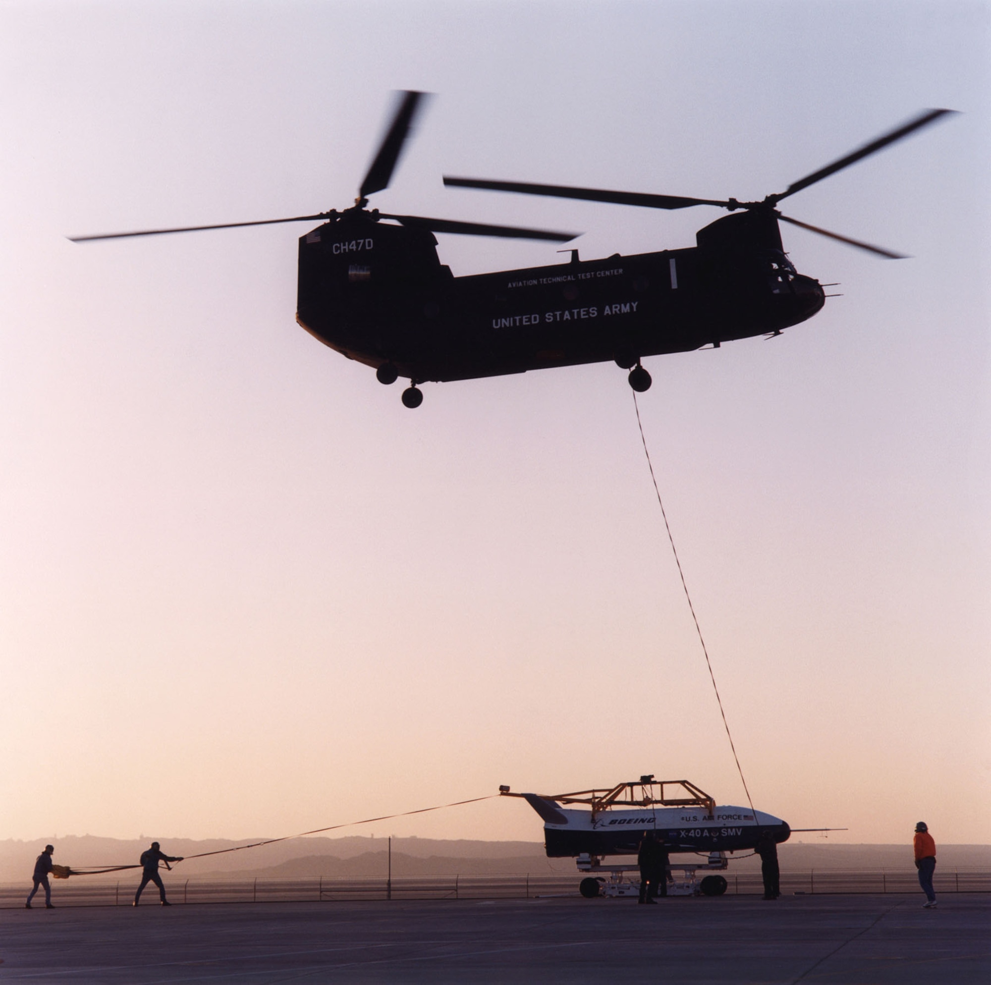 Boeing X-40A being lifted by a CH-47 helicopter for a free-fall test. Note the two support personnel holding the drag parachute behind the aircraft. Used to stabilize the X-40A while being lifted, the parachute was released just before the aircraft was released. (Photo courtesy of NASA)