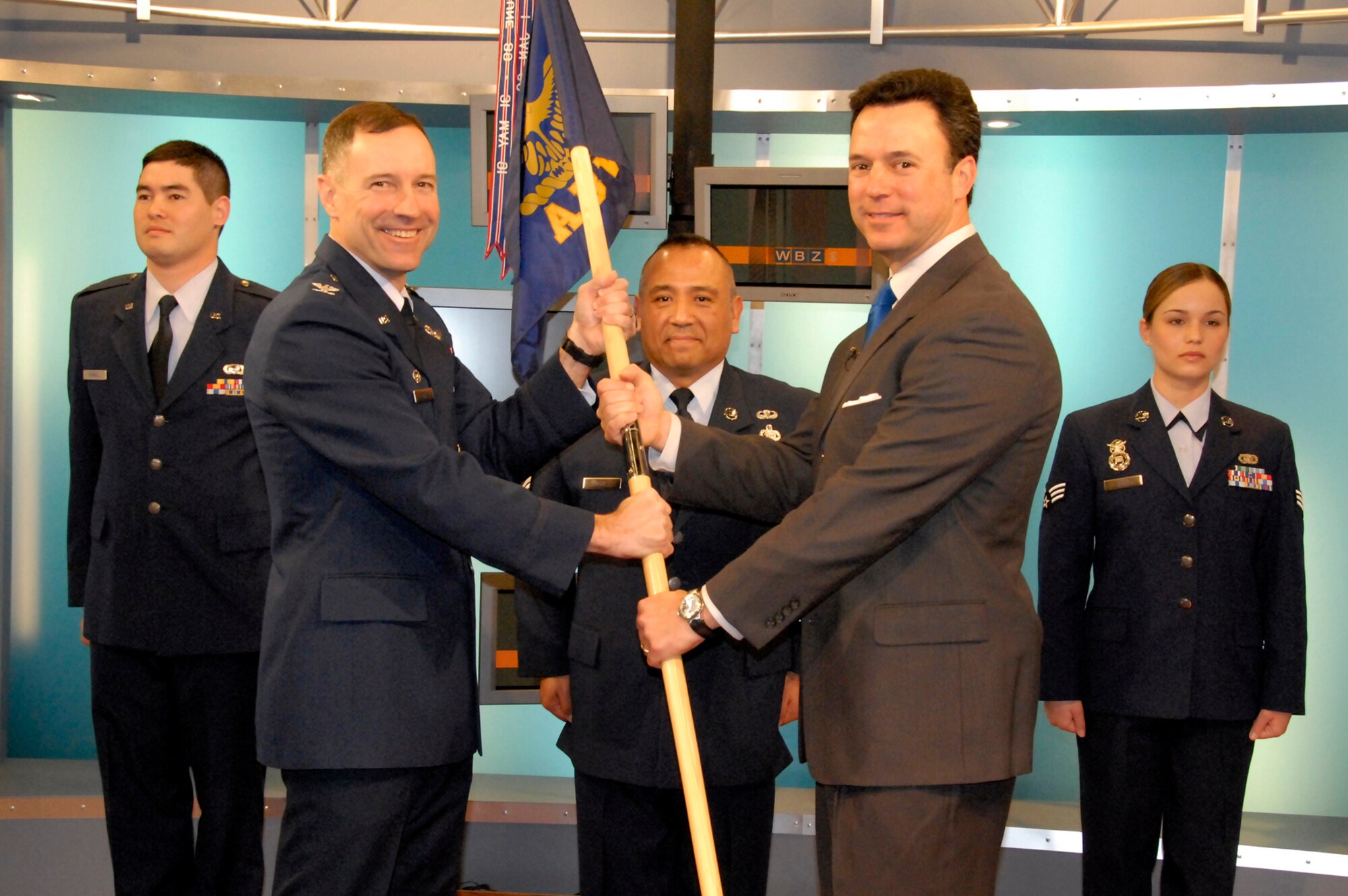 HANSCOM AIR FORCE BASE, Mass. -- WBZ-TV News Anchor Jonathan Elias (second from right) accepts the 66th Air Base Wing flag from 66 ABW Commander Col. David Orr during an Honorary Commander induction ceremony held Jan. 7, at the WBZ-TV station in Boston. First Lt. Will Russell, 66 ABW; Senior Master Sgt. Raul Ruiz, 66th Mission Support Squadron and Senior Airman Haley Roy, 66th Security Forces, participated in the ceremony. Additional Honorary Commanders will be inducted in a ceremony to be held Jan. 9 at 3 p.m. at the Minuteman Club. (U.S. Air Force photo by Mark Wyatt) 