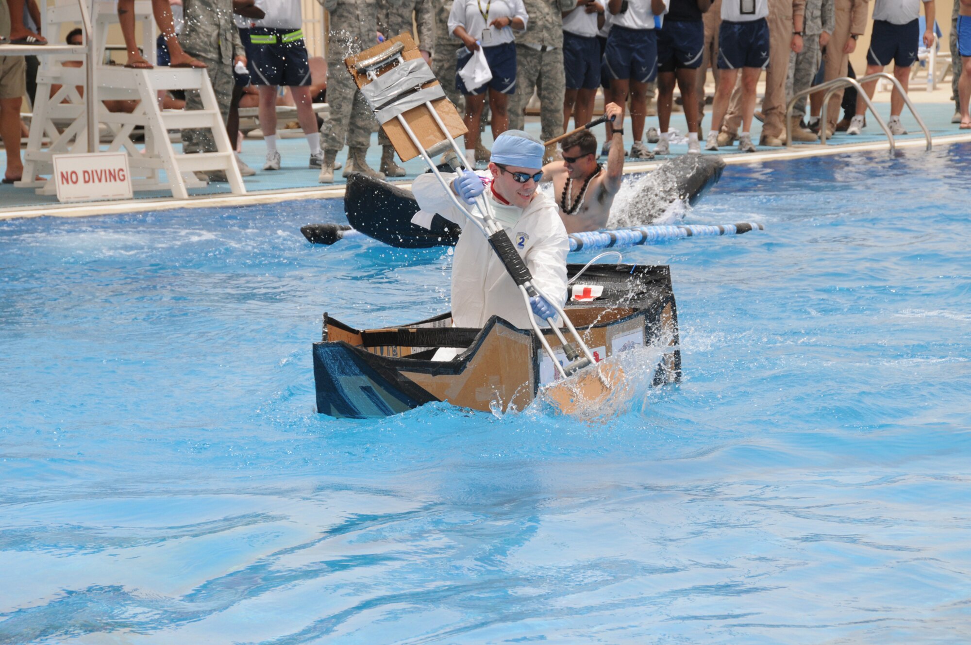 Senior Airman George Maceachern, 379th Expeditionary Medical Group, paddles across the swimming pool in the Cardboard Regatta during the Freedom Fest carnival. The 379th Expeditionary Maintenance Group won the event with their boat “Crash Recovery, EMX Pride of the Regatta.” (U.S. Air Force photo/Tech. Sgt. Shad Eidson)