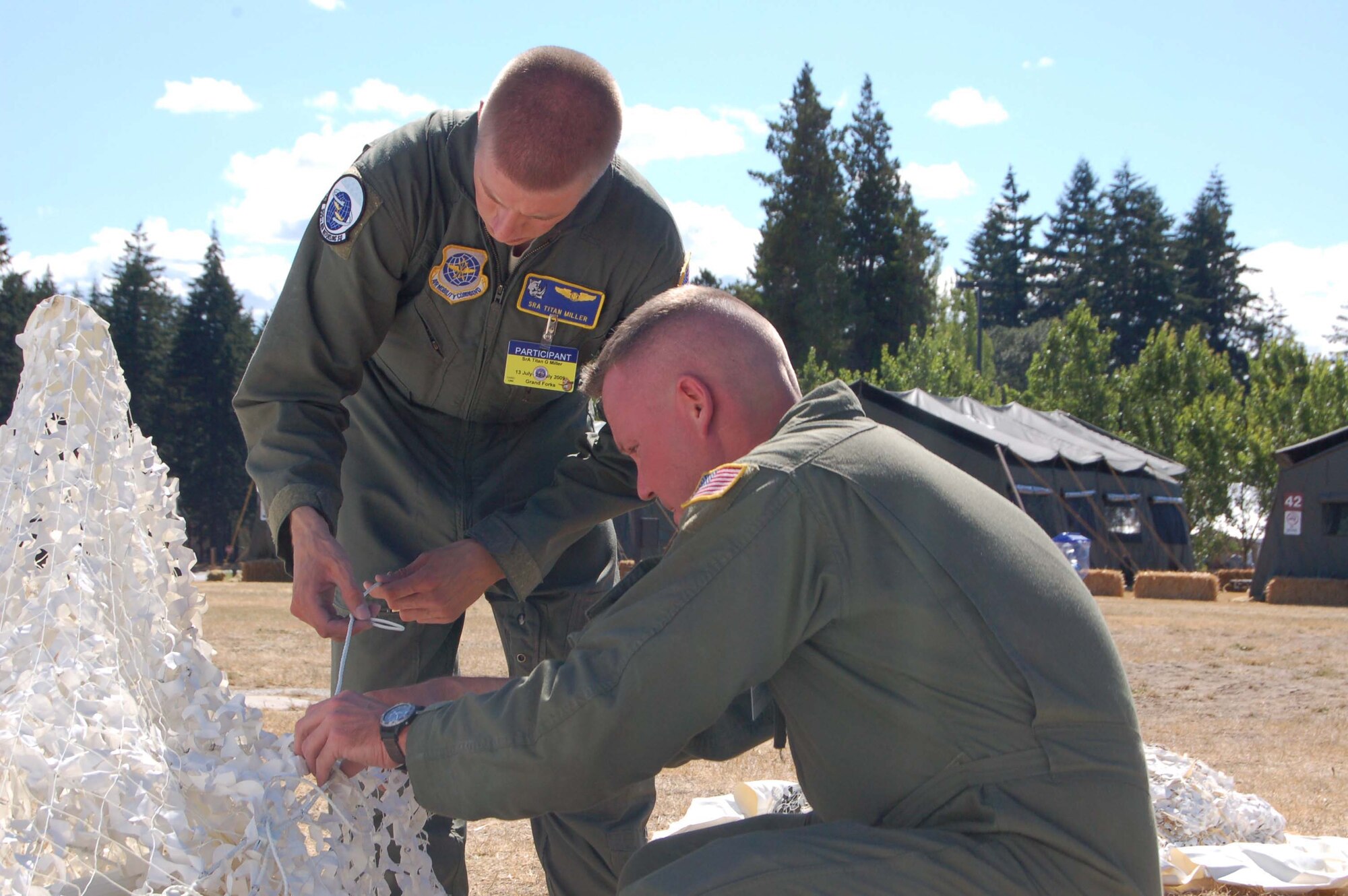 Senior Airman Titan Miller and Tech. Sgt. Izzy Hanks, both boom operators from the 905th Air Refueling Squadron, tie winter camo netting to a stake while setting up the hospitality tent in Renier Ranch at Air Mobility Command's Rodeo competition on McChord AFB, Wash., July 18.  (U.S. Air Force photo/Tech. Sgt. Amanda Callahan)