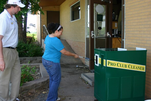 Jon Kula watches as Liz Choate washes a club in a new device the Tinker Golf Course has added to make life a little easier for players. The new Pro Washer is akin to a car wash for golf clubs. “By cleaning clubs and freeing the head from caked-on dirt it gives the club a better chance for optimal contact and greater backspin,” Kula said. The Pro Washer, a large green box located on the east side of the pro shop near the practice putting green, is free and simple to use. There is a green button on the front, push that and simply slide club in the washer to the left of the button. The washer can clean the grip end of the club as well. (Air Force photo by Kimberly Woodruff) 
