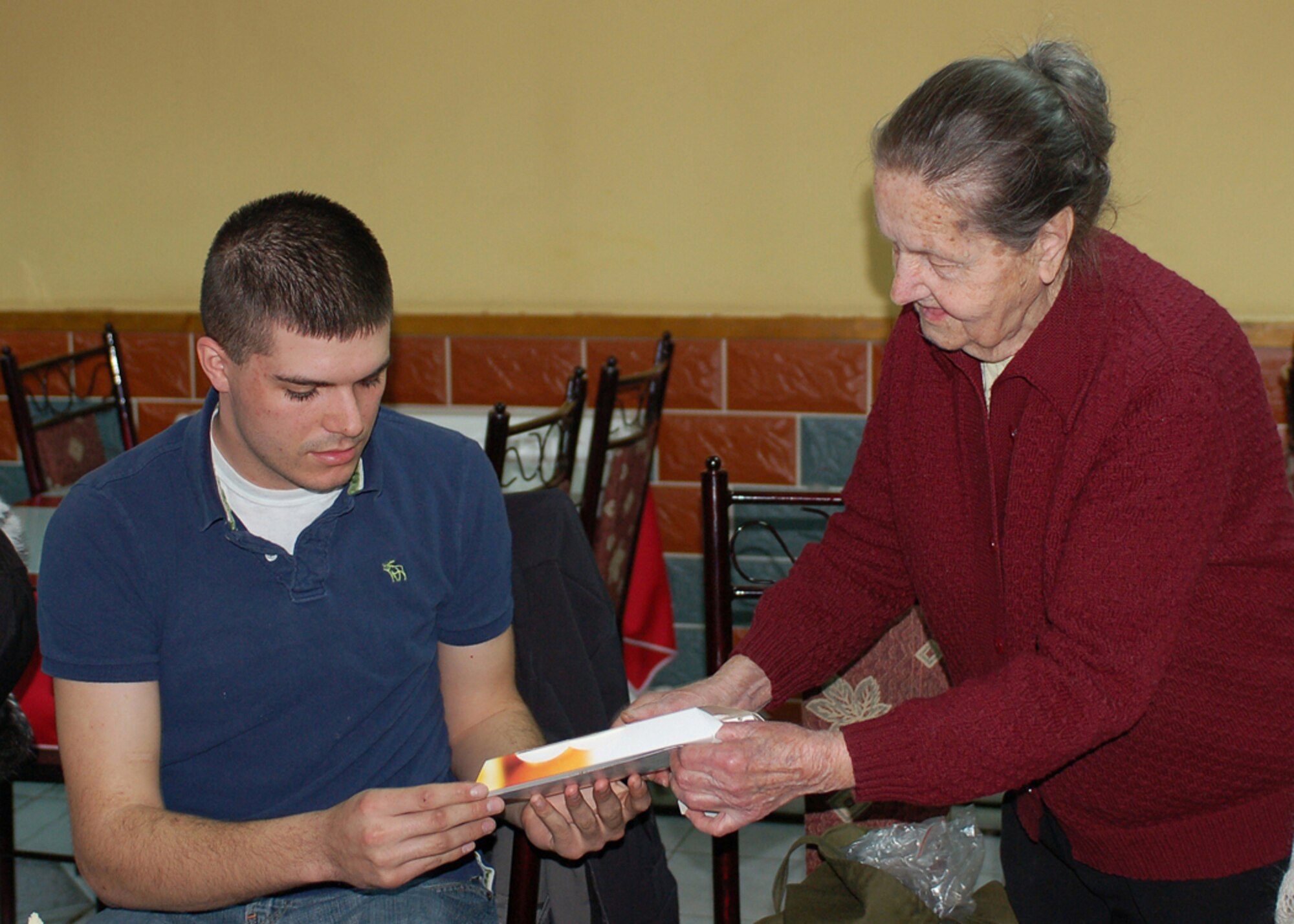 Airman 1st Class Jeremy Bowen helps Maria K. open a box of chocolates she brought to share with her friends from Manas Air Base who came to visit her and two other elderly ladies for lunch at a Turkish restaurant in Bishkek, Kyrgyzstan. Maria is known as a Babushka, or elderly woman, in Russian and Kyrgyz cultures. The 88-year-old woman has no family and was ?adopted,? along with two other elderly women, by the Manas Air Base Outreach Society through the Babushka Adoption Foundation, a local non-governmental organization. Each lady?s monthly income averages about 700 som or around $20 per month. Every month, volunteers from Manas join the ladies for lunch to share companionship, a little financial support and a small supply of groceries. Airman Bowen is assigned to the 22nd Expeditionary Air Refueling Squadron and is deployed from from the 92nd Operations Support Squadron at Fairchild Air Force Base, Wash. (U.S. Air Force photo/Tech. Sgt. Elizabeth Weinberg)
