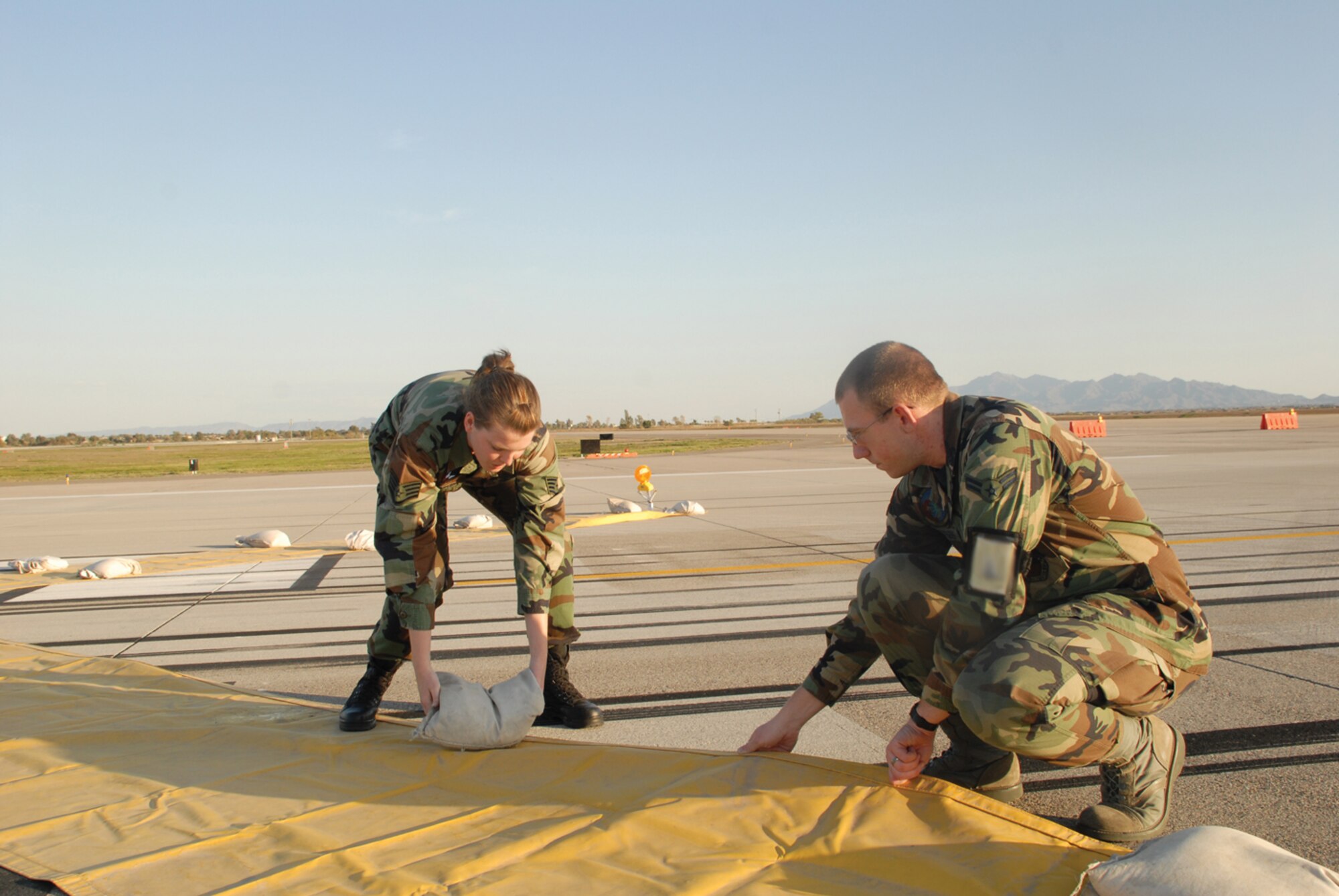 Staff Sgt. Kandace Parker, 56th Operations Support Squadron airfield management operations supervisor, and Airman 1st Class Lawrence Stephens, 56th OSS airfield management operations coordinator, readjust sand bags on a marker on the Luke Air Force Base flightline. The marker, a large yellow “X,” is used to warn pilots of a runway closure. (U.S. Air Force photo/ Airman 1st Class Tracie Forte.)