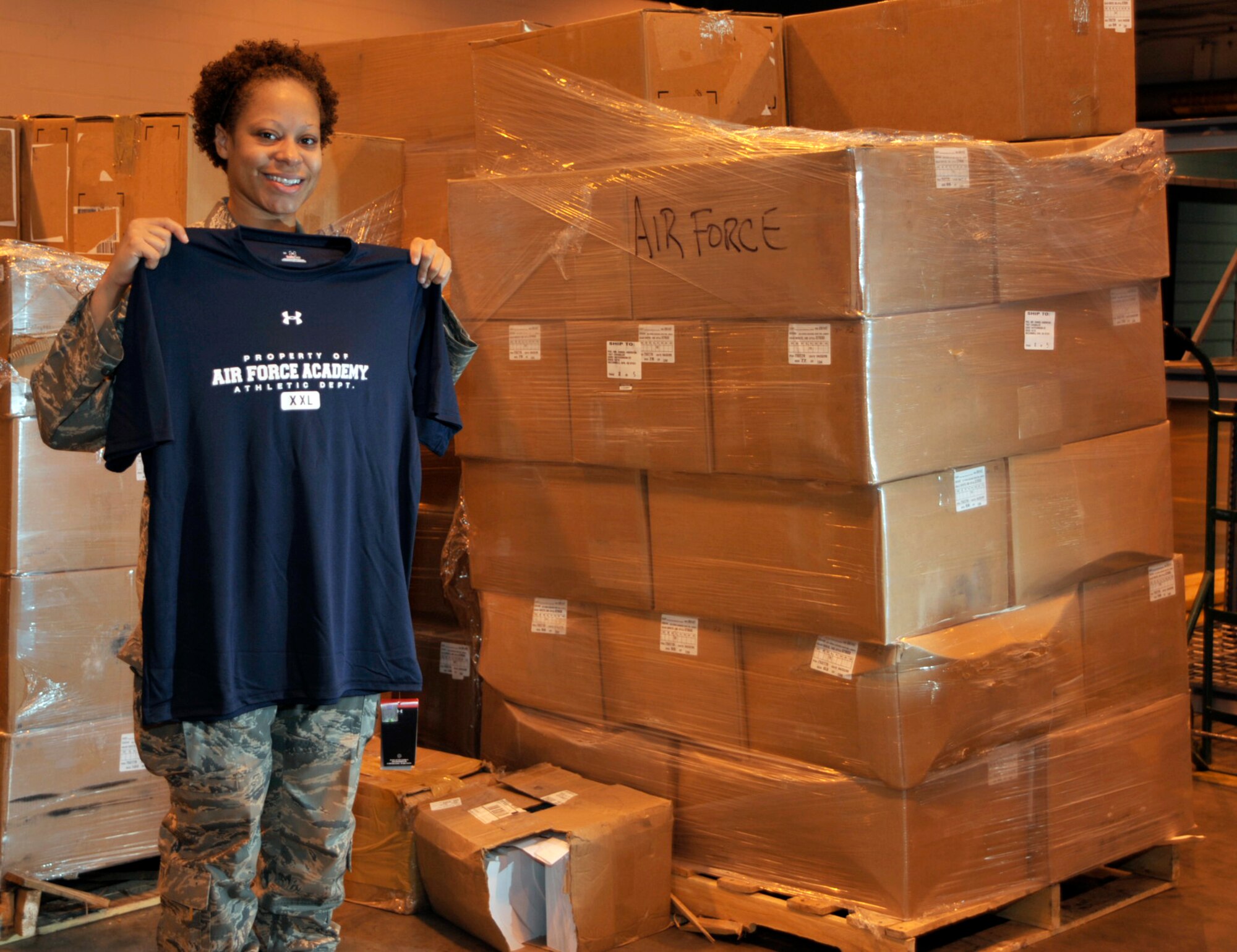 Staff Sgt. Tina Nero stands with one of the many T-shirts filling her warehouse at McConnell Air Force Base, Kan. Sergeant Nero is part of a 22nd Logistics Readiness Squadron team at McConnell processing a large amount of cargo for Coaches Tour 2009, a morale-boosting mission designed to bring NCAA football coaches to troops overseas. The shirt Sergeant Nero holds was donated by Under Armour, the main sponsor of Coaches Tour. Air Force head coach Troy Calhoun, one of seven coaches slated for the tour, will hand out many of the shirts in the boxes behind Sergeant Nero labelled "Air Force."