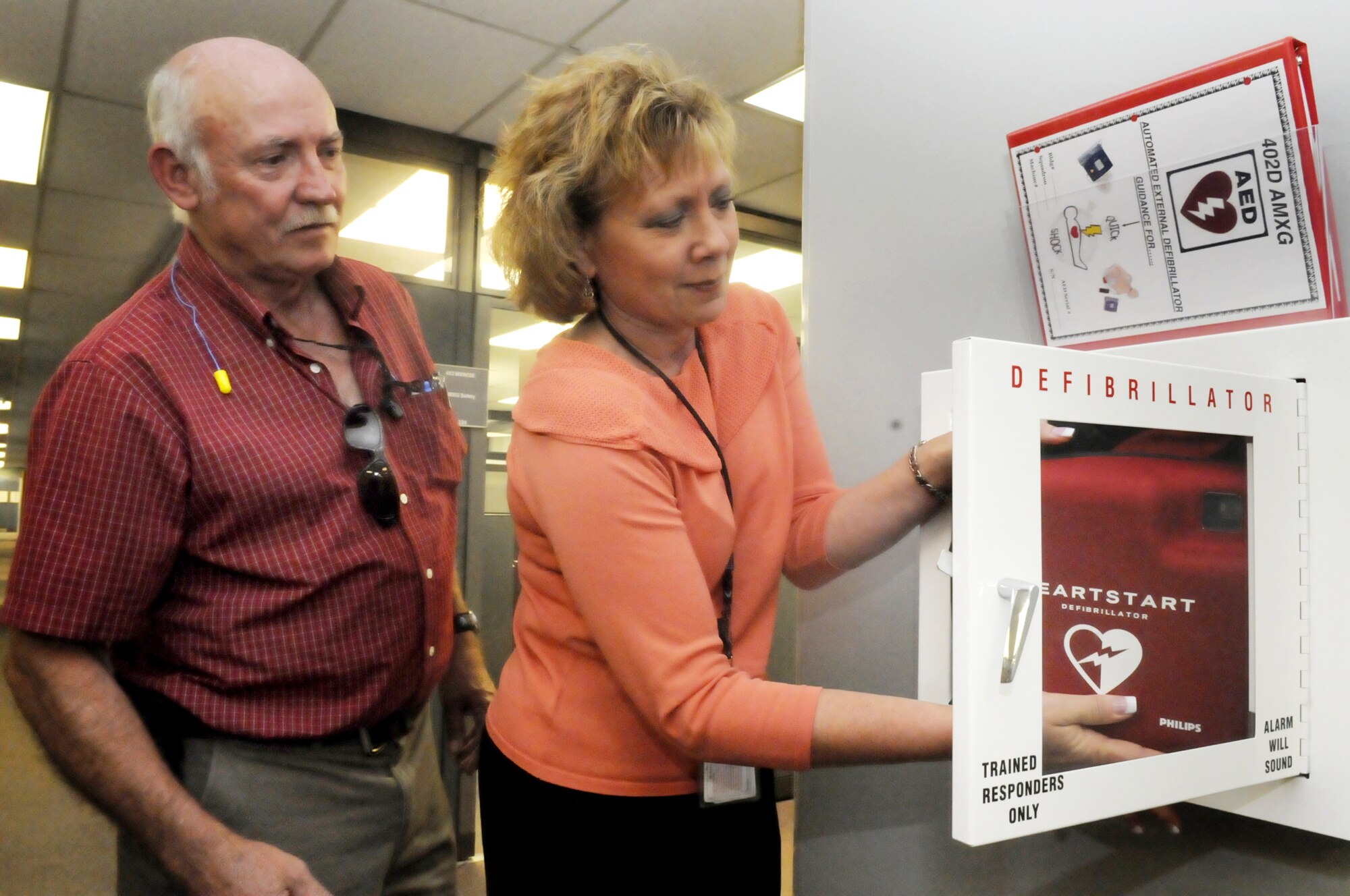 David Coody, owner of Coody Electric, which is installing all of the defibrillator wall cases in the flightline area, watches as Linda King, Commander’s Action Group executive program management support specialist, puts one of the new defibrillators in place in the hallway of Bldg. 125. U. S. Air Force photo by Sue Sapp
